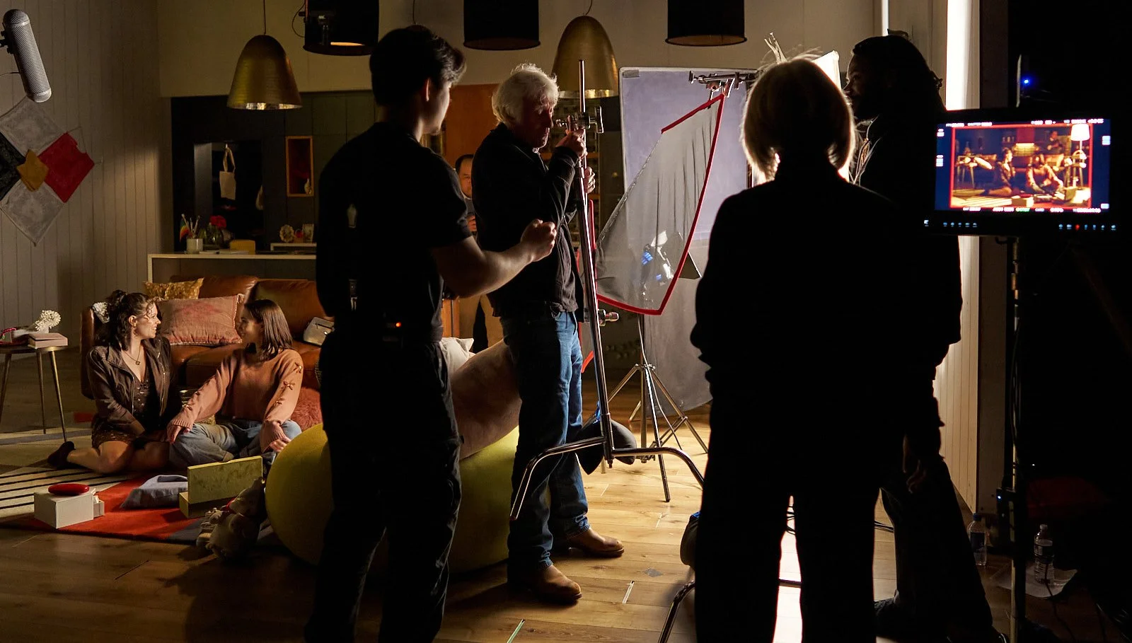 Film crew filming a scene indoors with two women sitting on the floor and three people operating equipment and cameras.