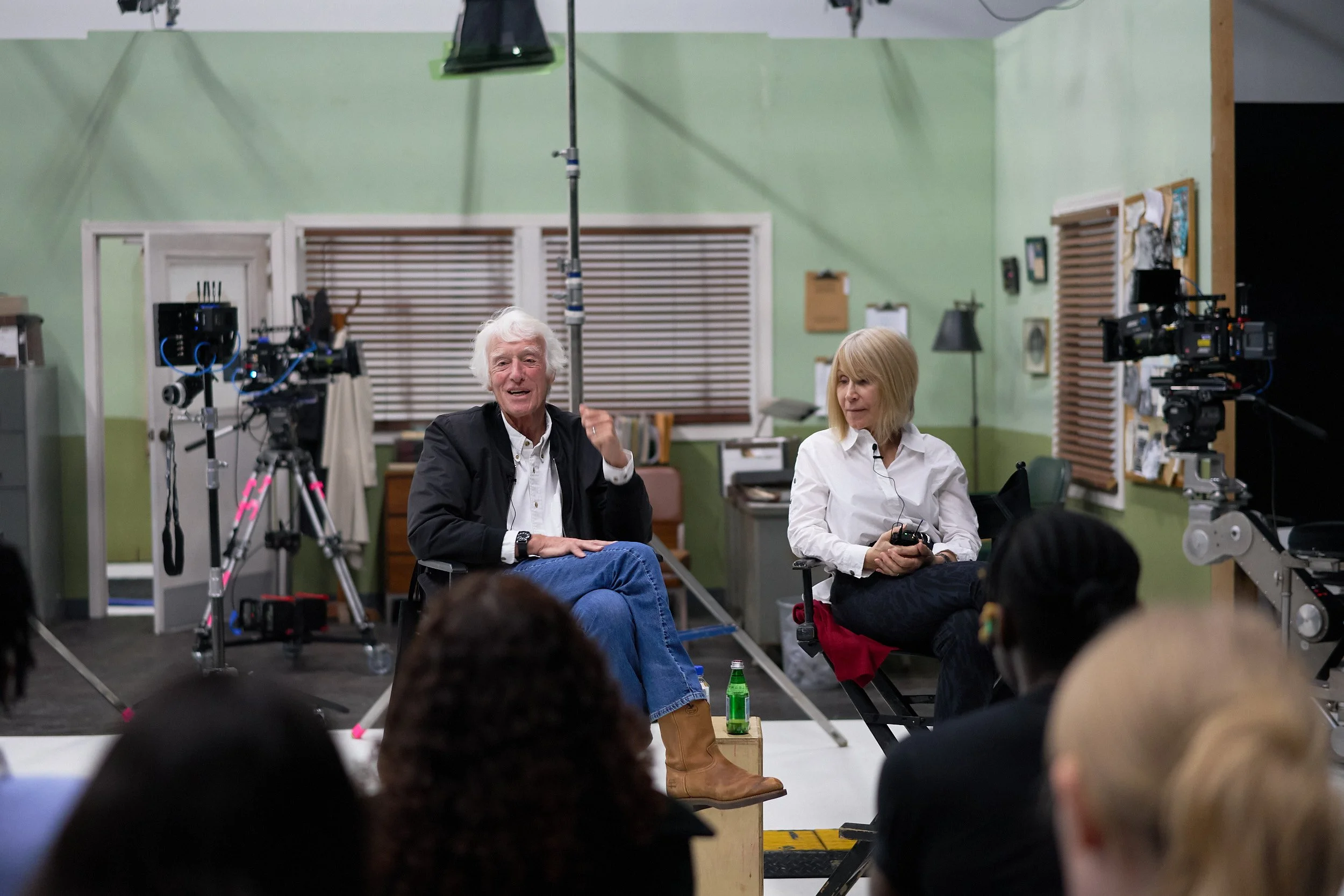 Two people, a man with white hair and a woman with blonde hair, are seated and speaking during a filming or video recording session in a green-colored room with cameras and filming equipment around them. The man is wearing a black blazer, white shirt