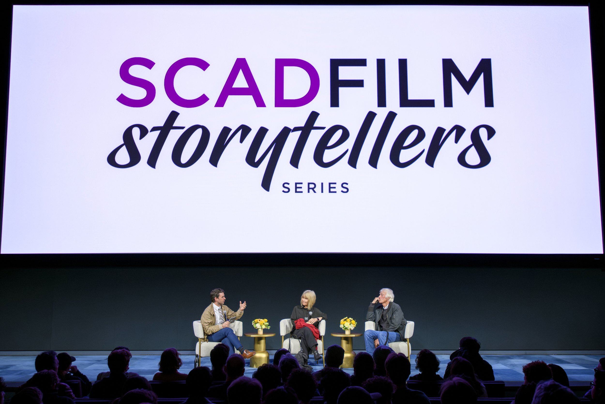 A panel discussion at a conference or event with three people seated on stage, engaged in conversation, with microphones and small tables with flower arrangements in front of them. A large screen above displays the event name 'SCADFILM storytellers S