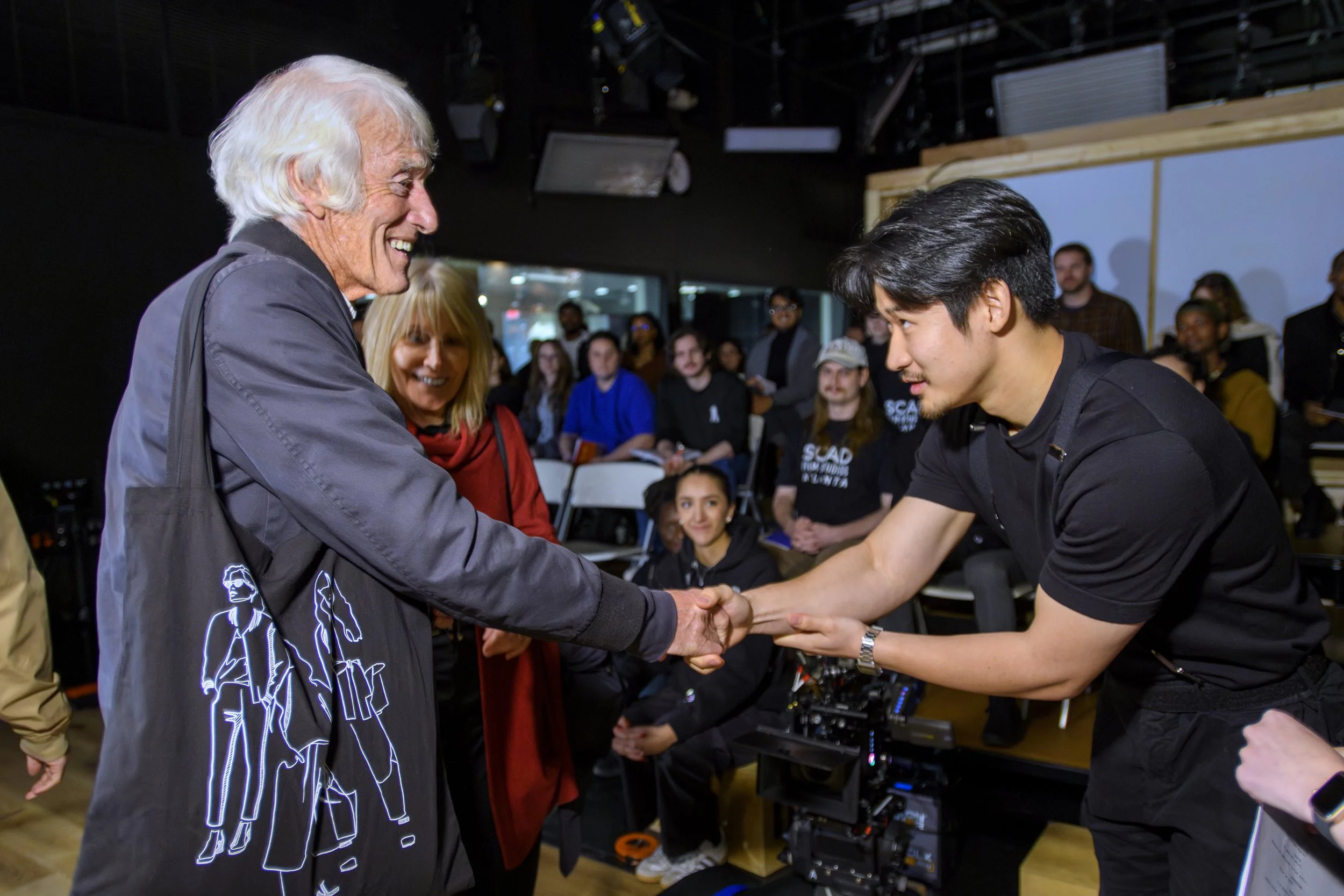 An elderly man with white hair wearing a black jacket shaking hands with a young man in black shirt, with smiling crowd in the background.