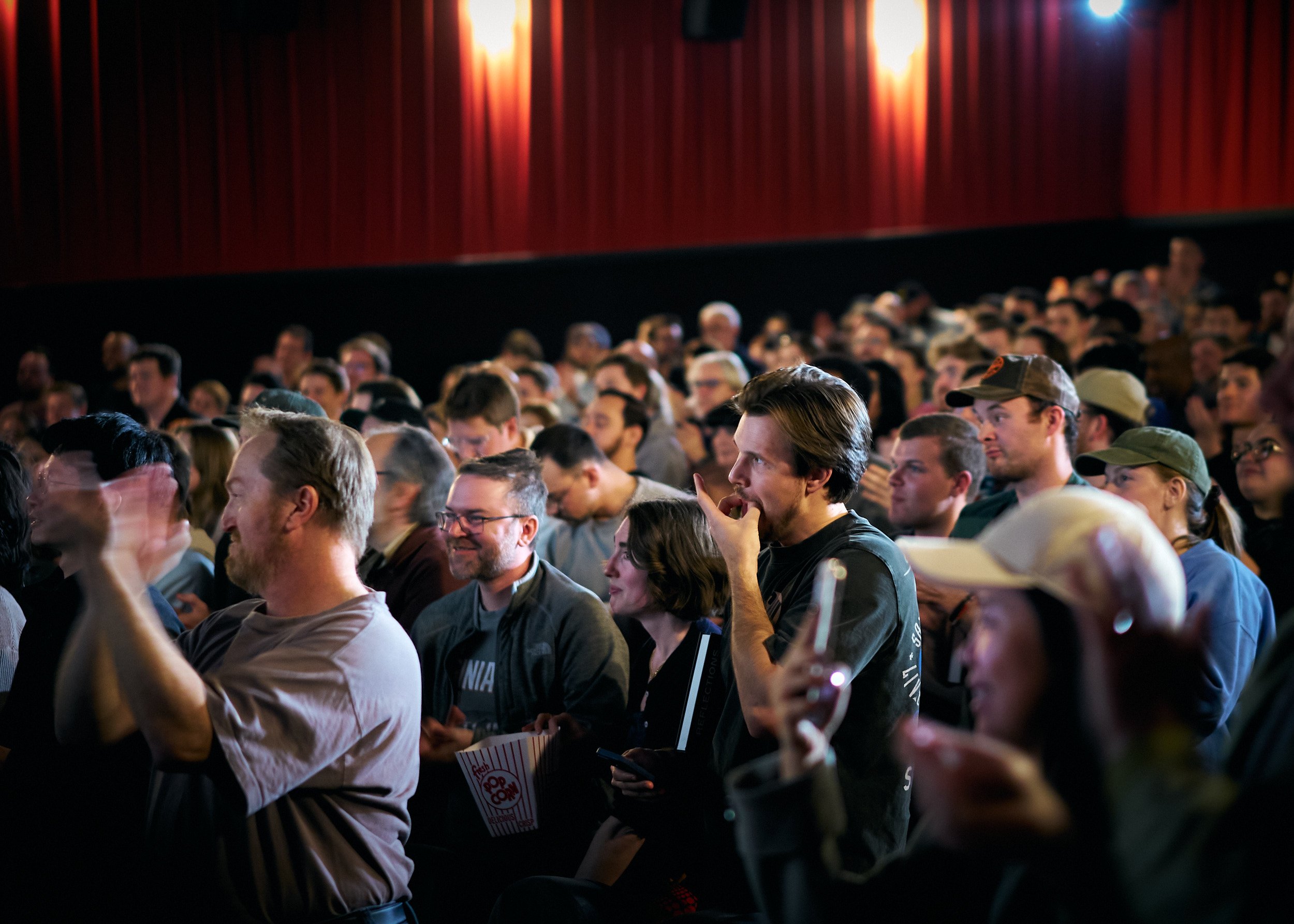 Crowd of people seated in a theater with red curtains, some are looking at their phones, some are talking, and others are watching the stage.