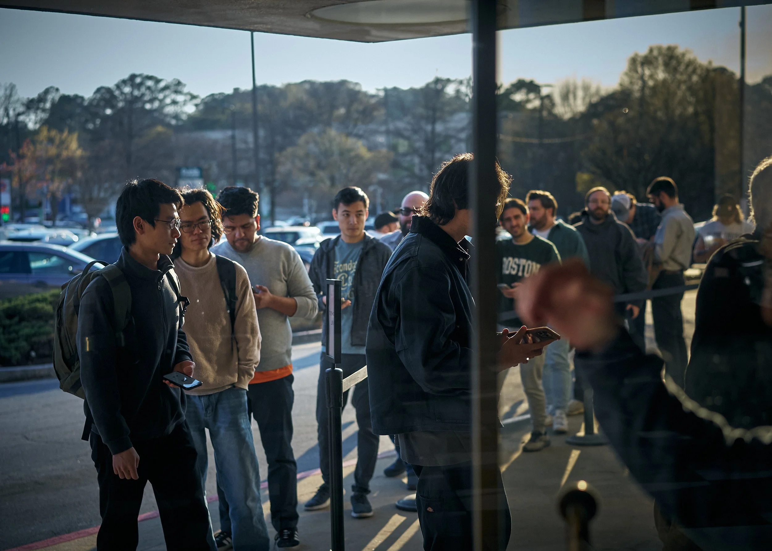 People standing in line outdoors during daytime, waiting to enter a building with trees and parked cars in the background.