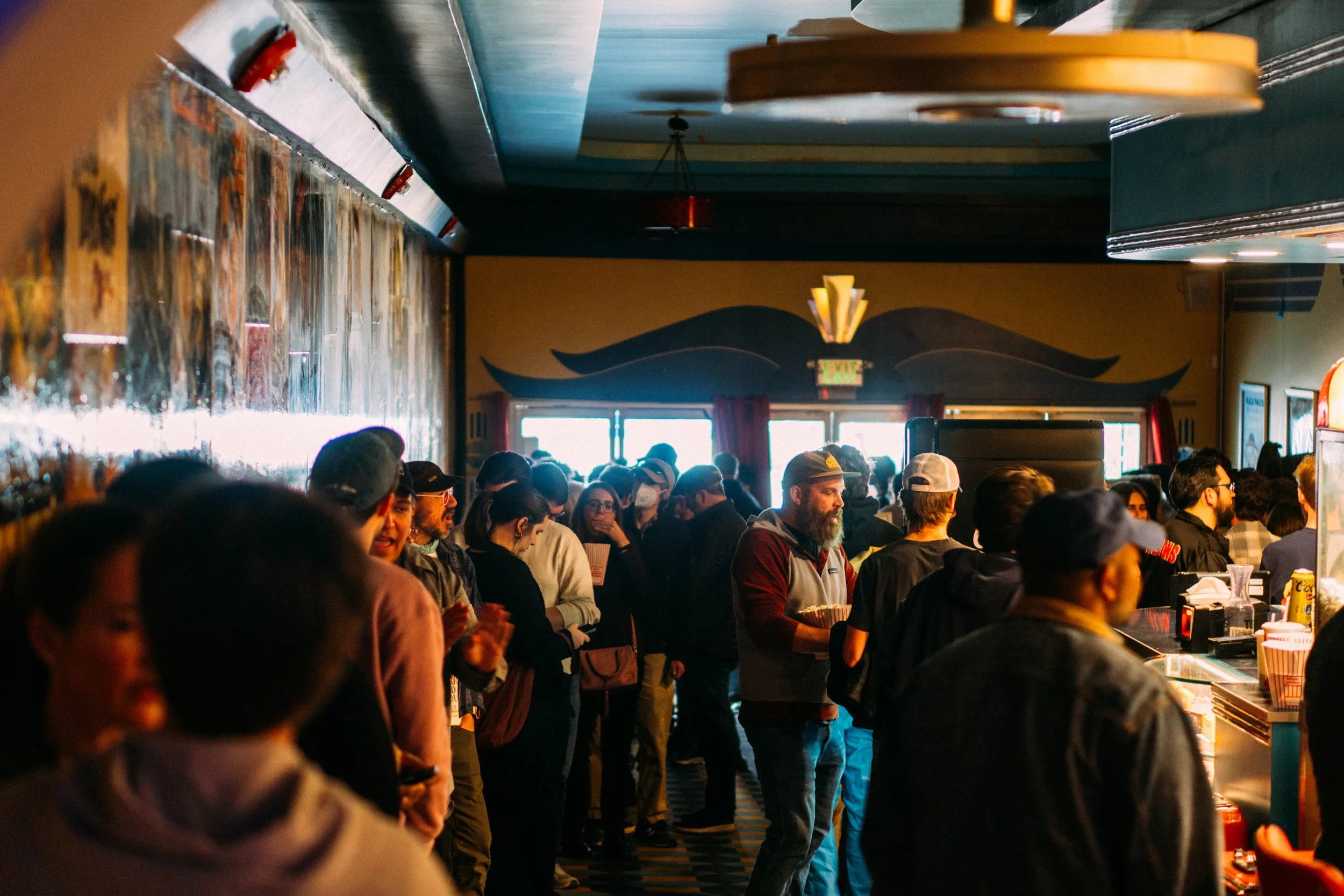 Crowd of people inside a restaurant or bar, with some standing at a counter, with colorful walls and a large window in the background.