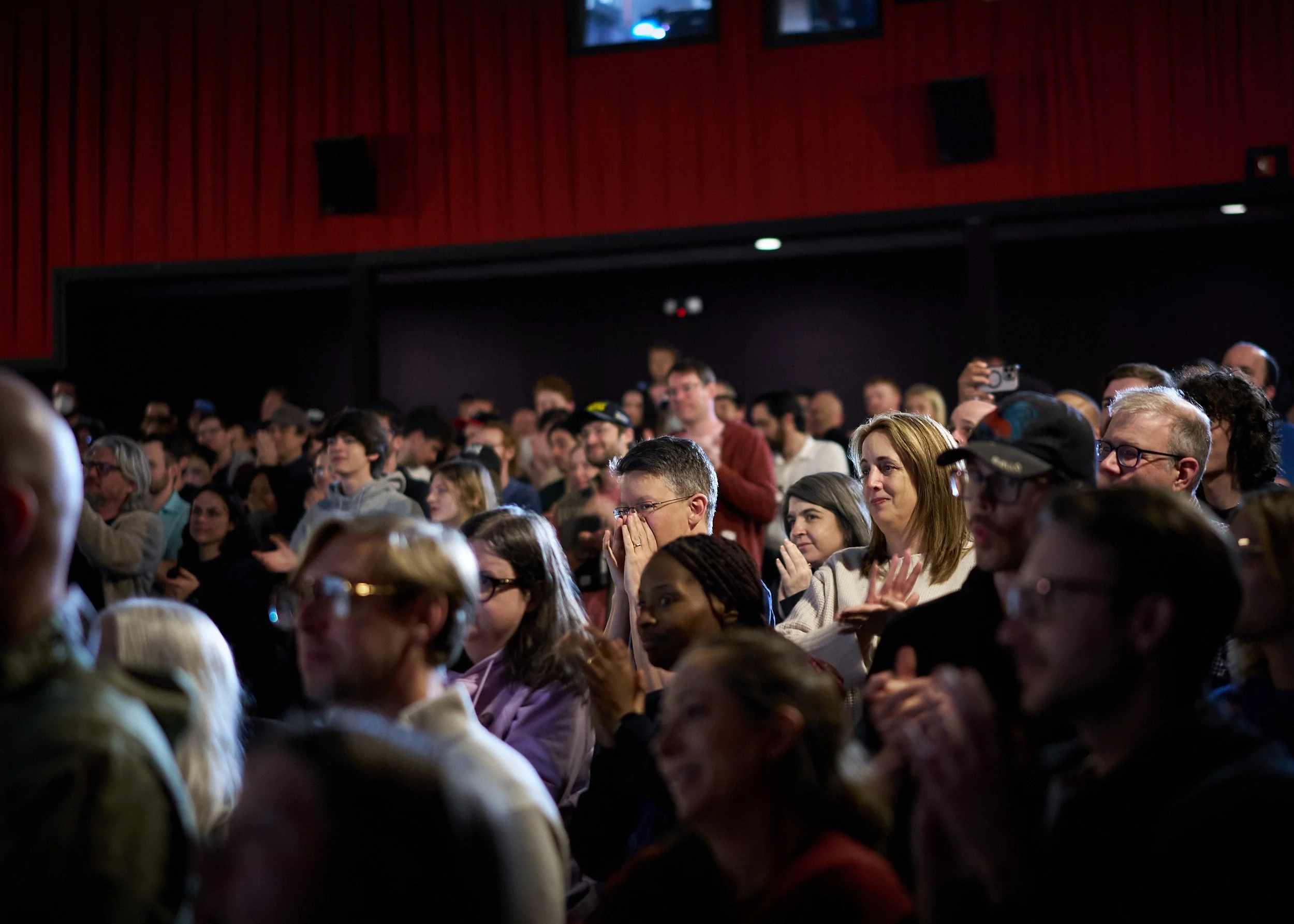 A large group of people gathered in an indoor space, some are standing and some are sitting, with a focus on a few individuals in the foreground and others in the background, with red wall paneling and mounted screens.