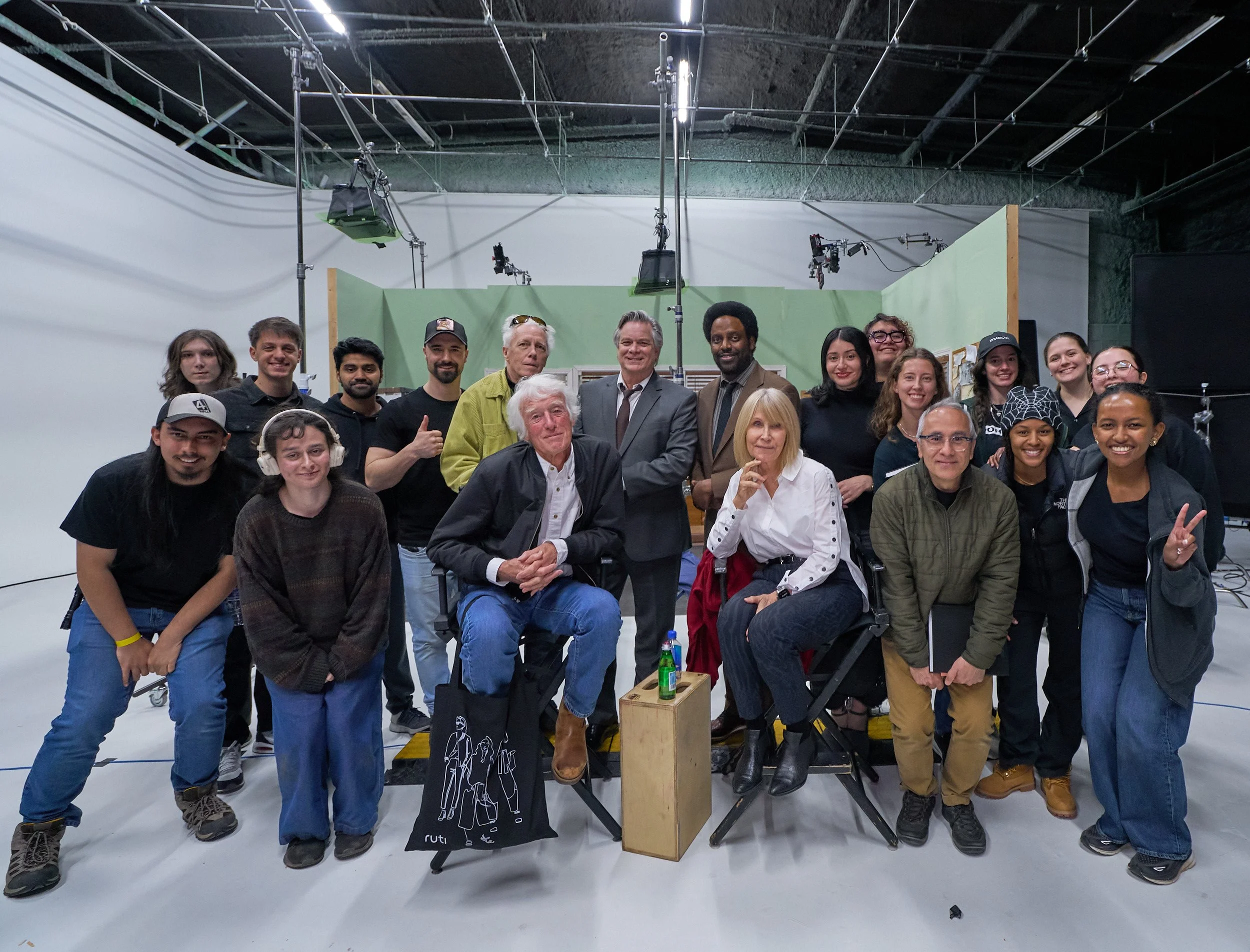 A group of diverse people gathered in front of a green screen with filmmaking and lighting equipment overhead. They are posing for a photo on a film set.
