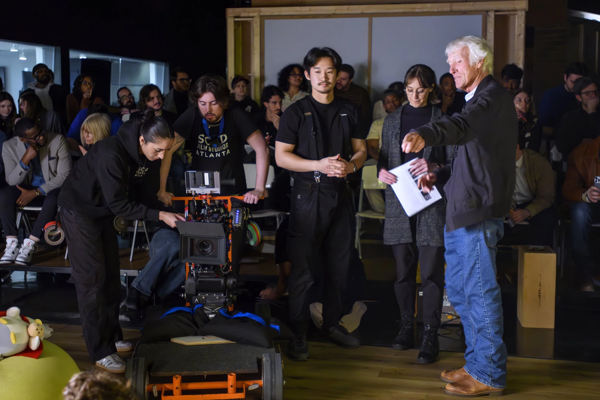 Group of people, including camera crew and audience, gathered around a person working on filming a scene with a professional camera in an indoor setting with wooden and glass walls.