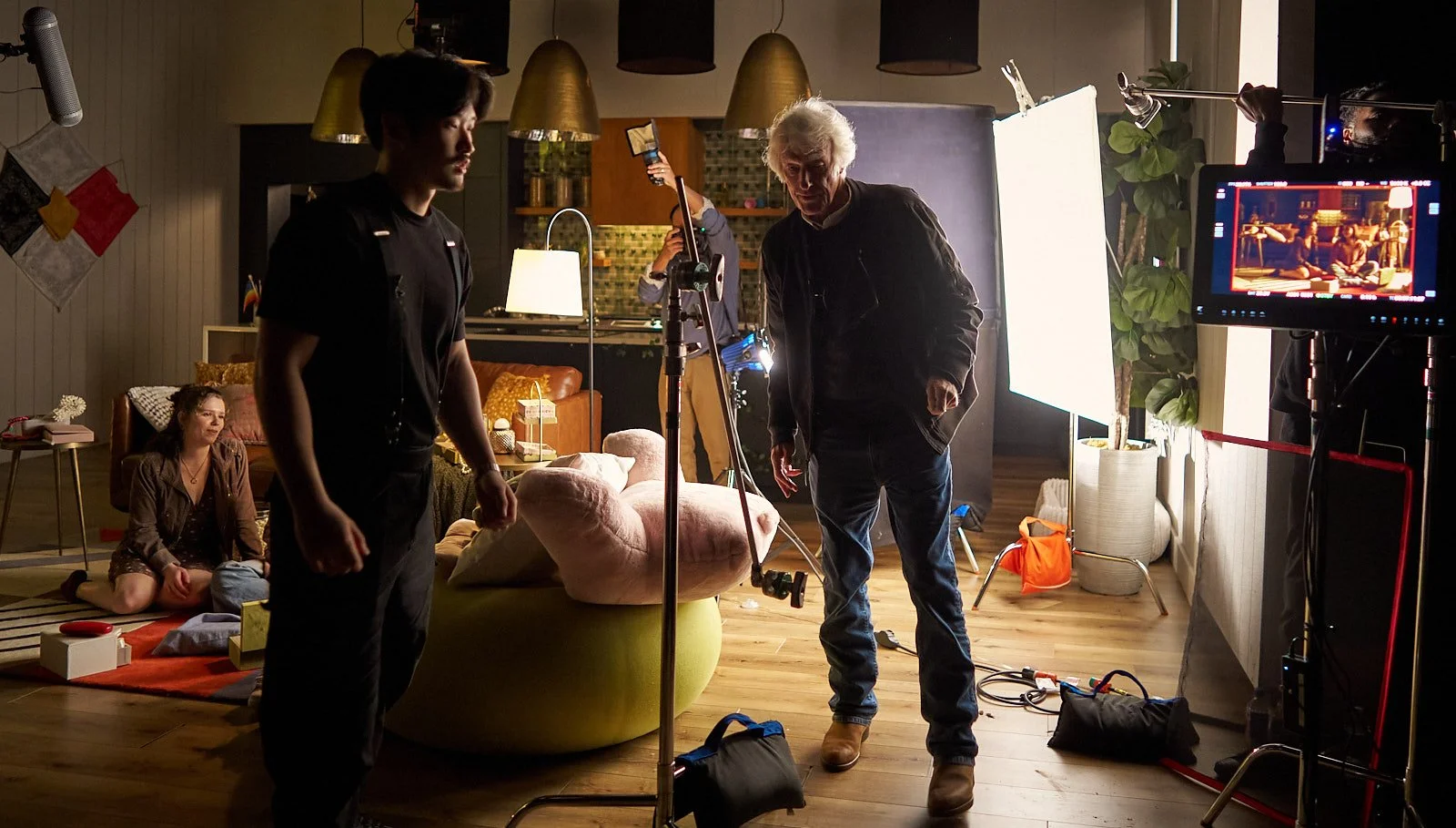 Filming a scene in a cozy living room with two women sitting on the floor, a man in dark clothing and an older man with white hair standing near filming equipment, camera monitors, and studio lights.