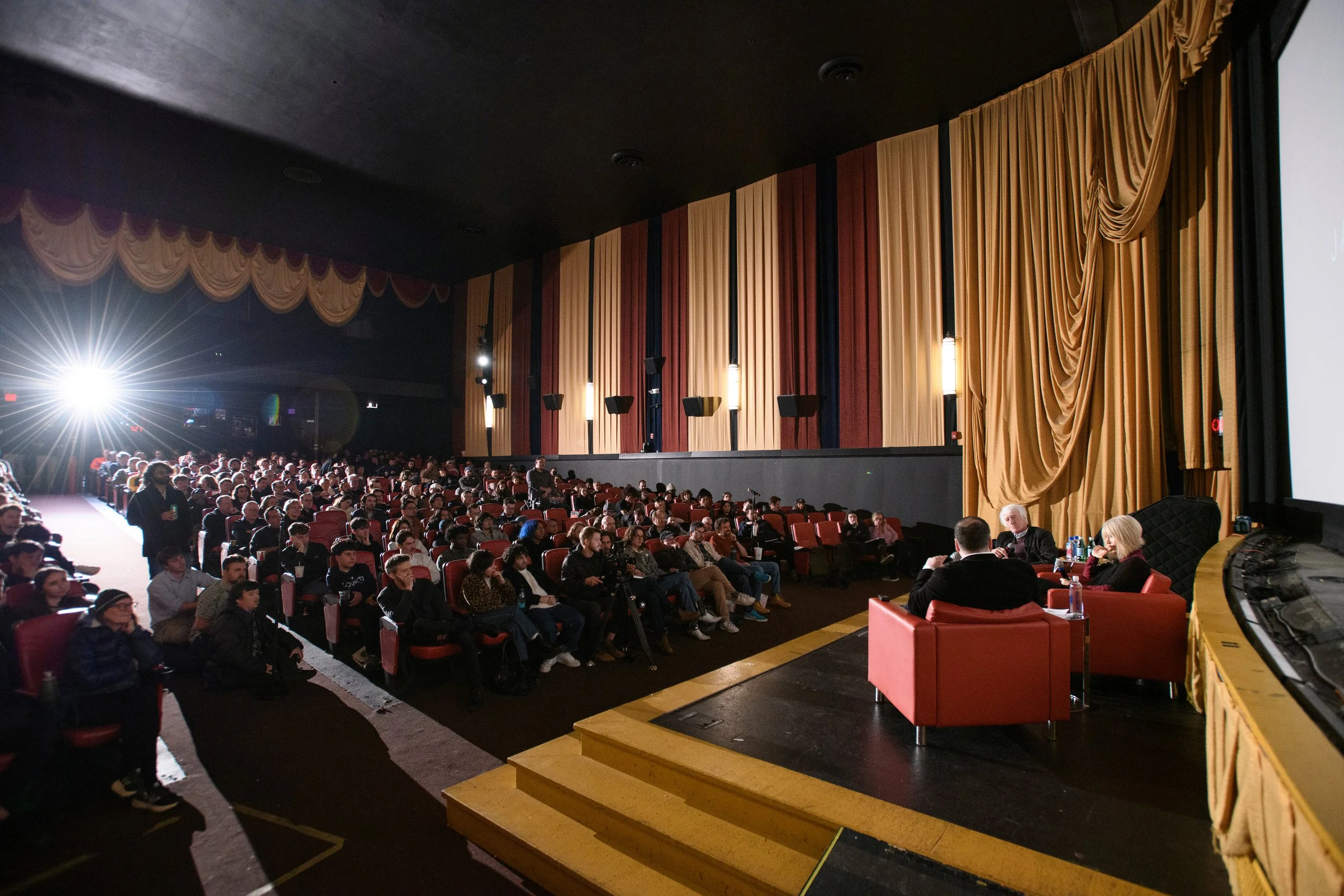 Audience seated in theater listening to presentation on stage with curtains