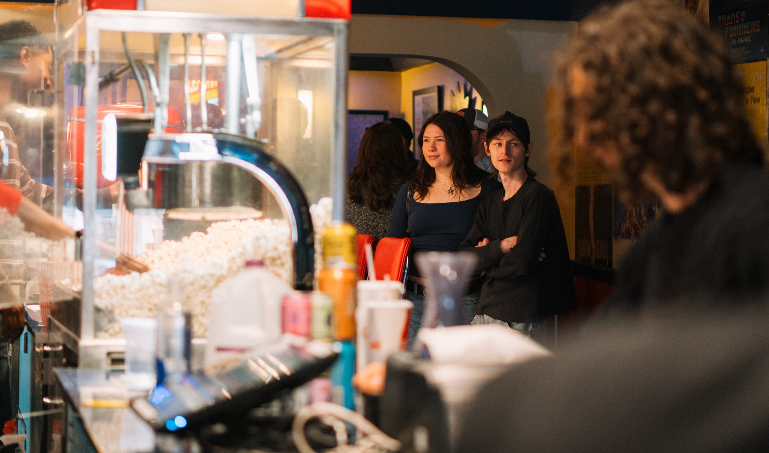 People standing in line inside a movie theater concession area, with popcorn and snacks visible in the foreground.