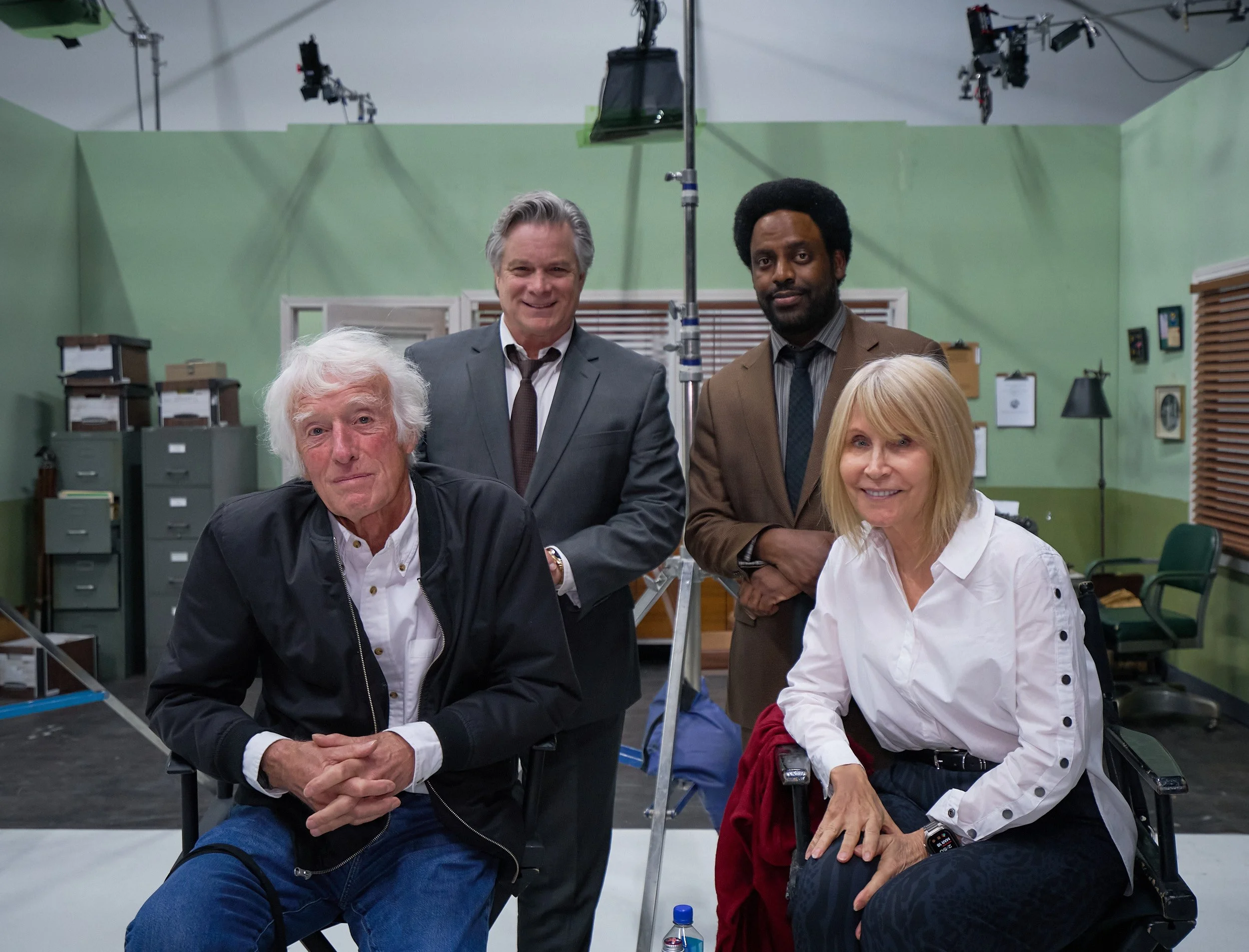 Group of five people, three women and two men, gathered in a room with green walls, standing around camera equipment and a green screen background, possibly on a film set.