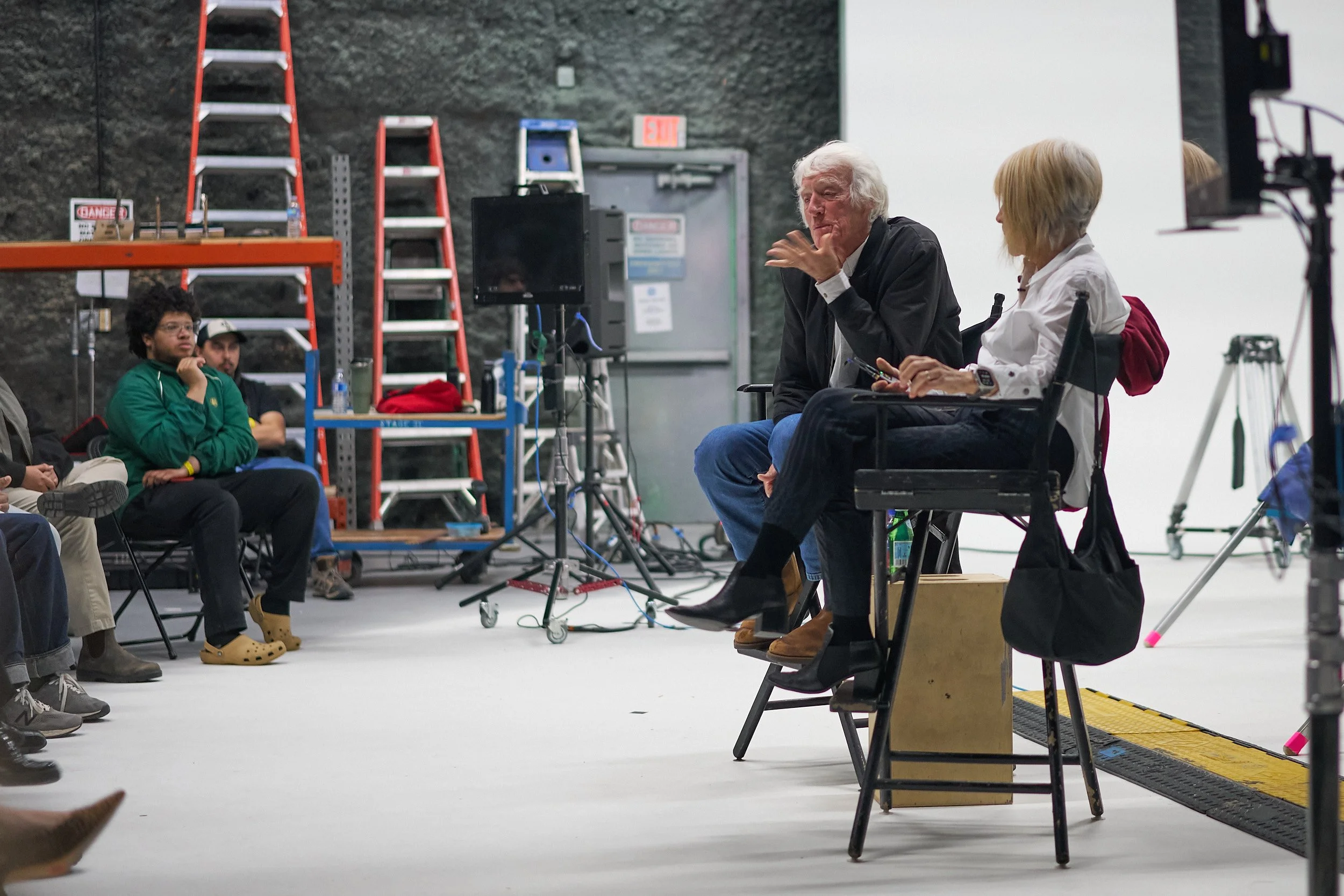 A group of people sitting in a studio or workshop. An older man with white hair is talking while seated, facing a woman with blonde hair who is taking notes. In the background, there are ladders, shelves, and technical equipment.