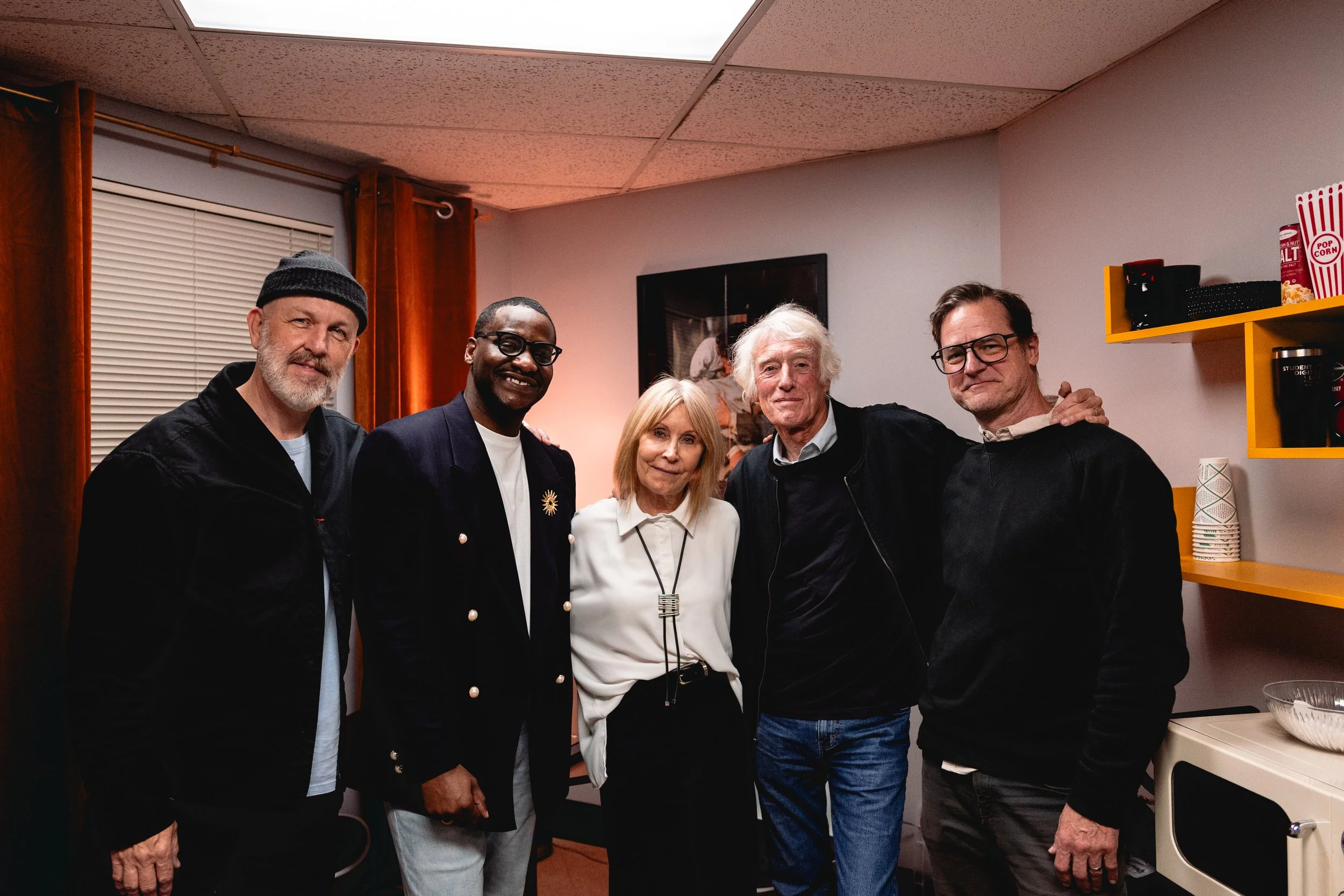 Group of five people standing together in a room, smiling for the photo. There are three men and two women, with a mixture of ages and appearances.