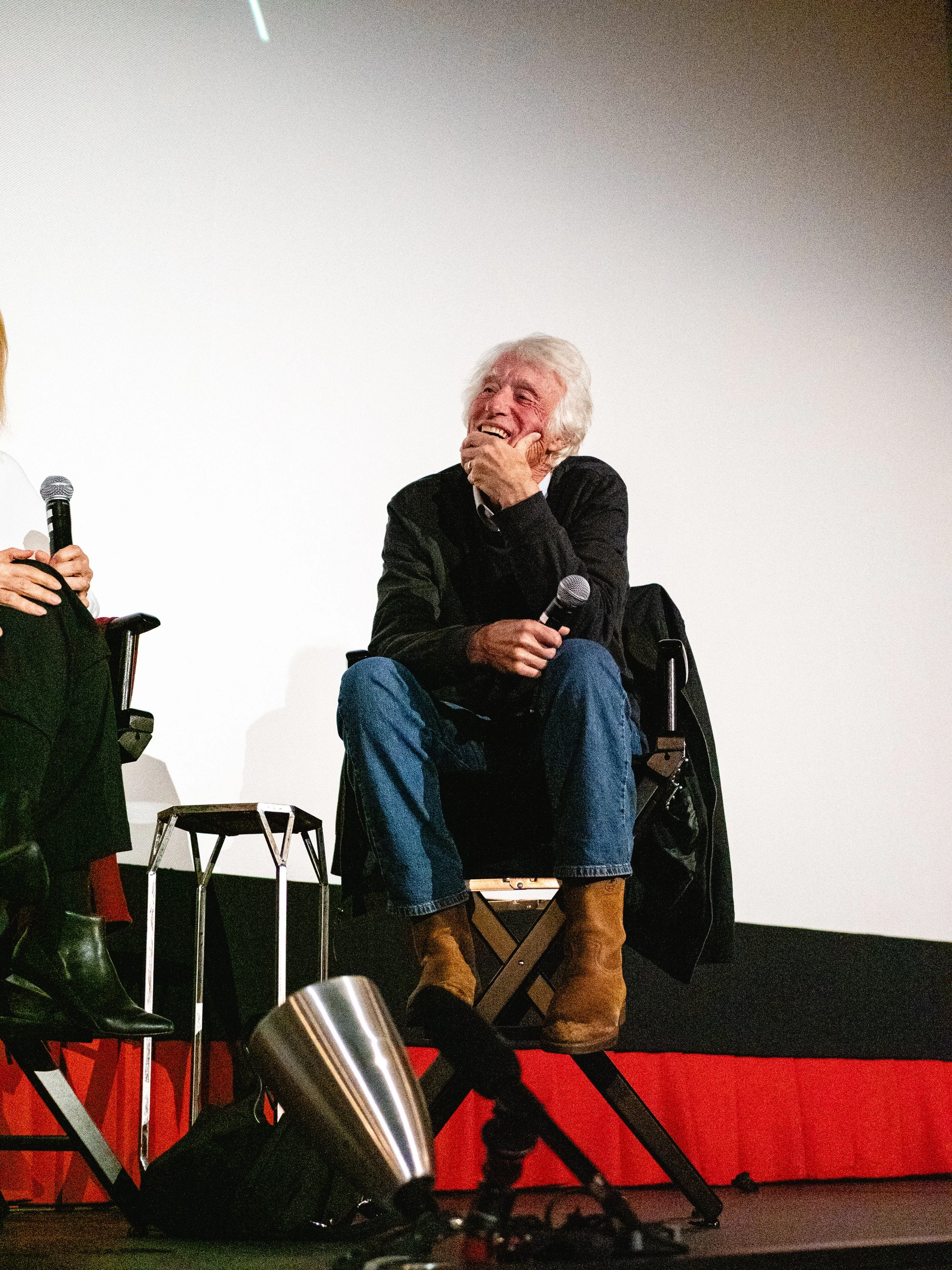 An elderly man with white hair, dressed in jeans, a black jacket, and brown boots, sitting in a wheelchair on a stage, holding a microphone in his right hand, and laughing while looking to his left. There are microphones and a laptop on a table besid