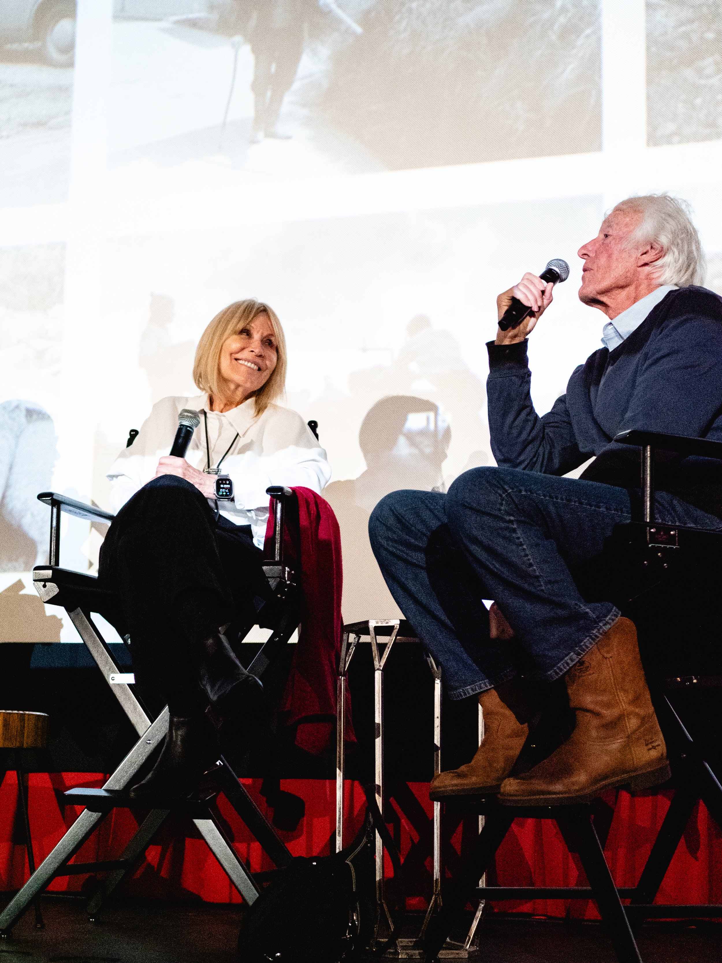 An elderly man and woman sitting on stage during a panel discussion or interview, both holding microphones. The woman is smiling and looking at the man. The background features a large screen displaying part of the stage and some audience members.