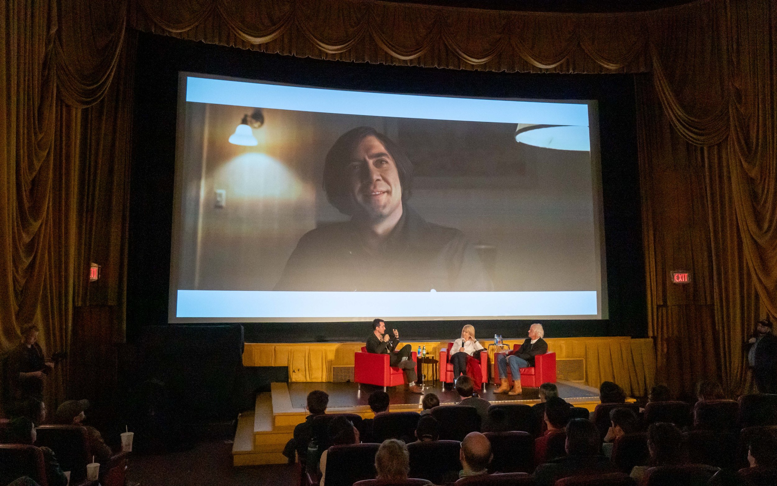 An auditorium with a large screen showing a man's face. There are multiple people seated on stage, including a person in a dark blazer speaking into a microphone, a woman in a white top, and an older man in a dark suit. Audience members are sitting i
