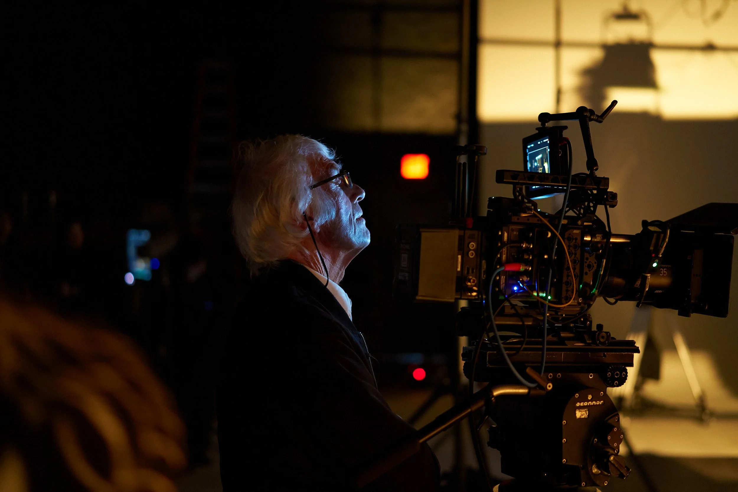A woman with white hair and glasses is operating a professional camera on a film set, with a brightly lit screen and equipment around her in a darkened environment.
