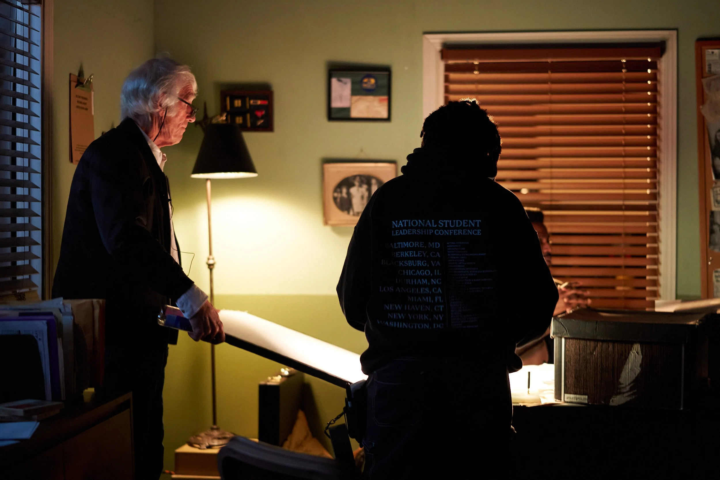 Two people are in a dimly lit room with a green wall, engaged in a conversation. One older man with white hair and glasses is on the left, and a person with short curly hair wearing a black hoodie with references to multiple cities is on the right. T