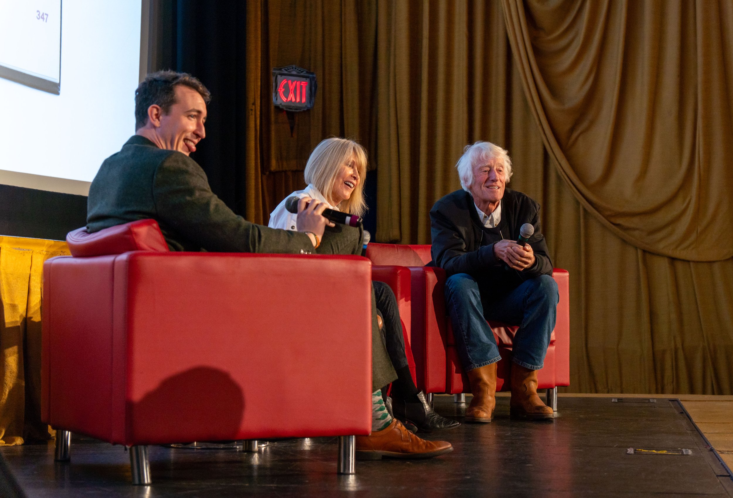 Three people sitting on red chairs on a stage during a panel discussion or interview, with a woman holding a microphone, a man smiling, and an older man also holding a microphone. There is a curtain in the background and a projection screen to the le
