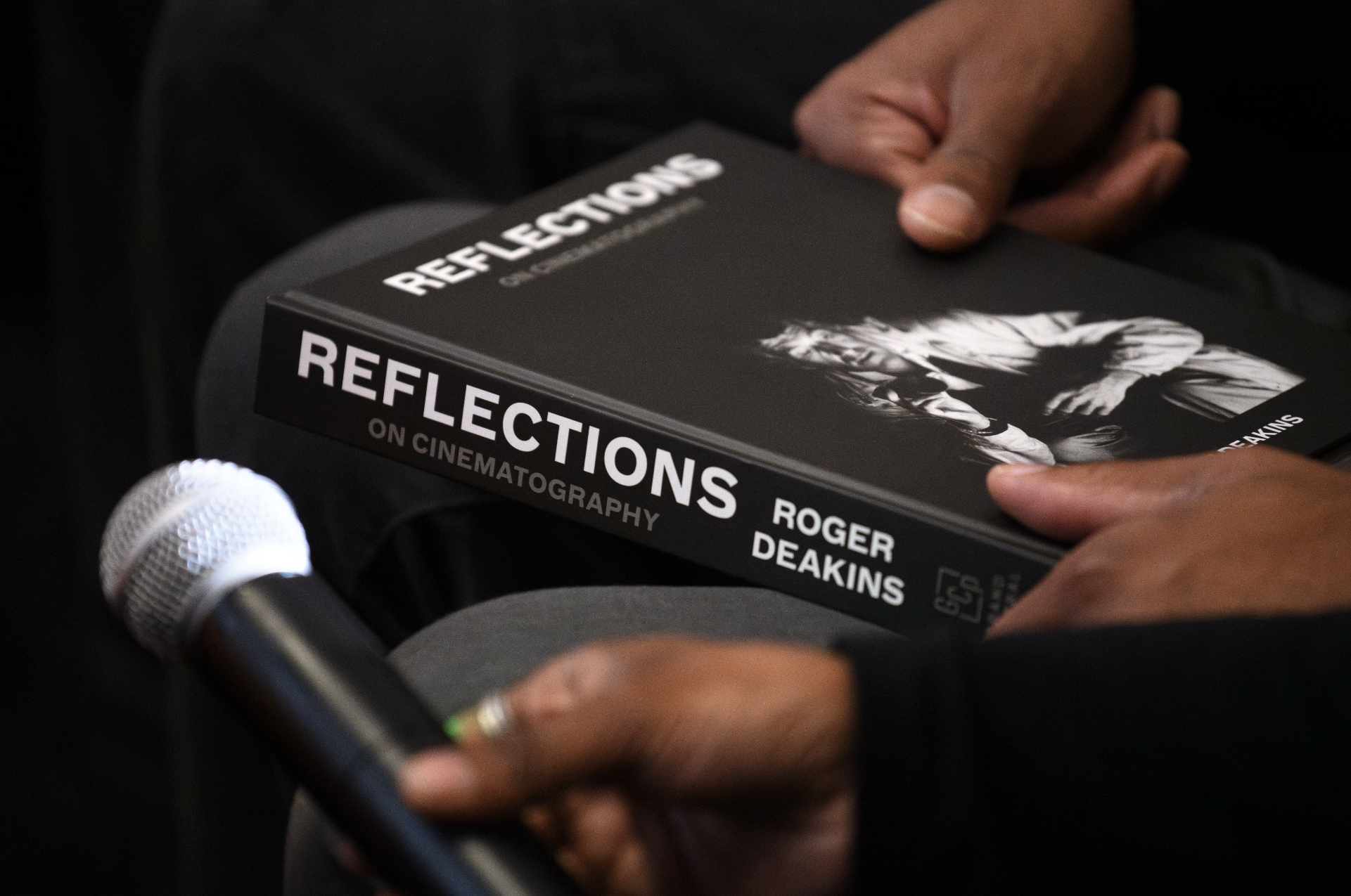 A person holding a book titled 'Reflections on Cinematography' by Roger Deakins, with a microphone in the foreground.