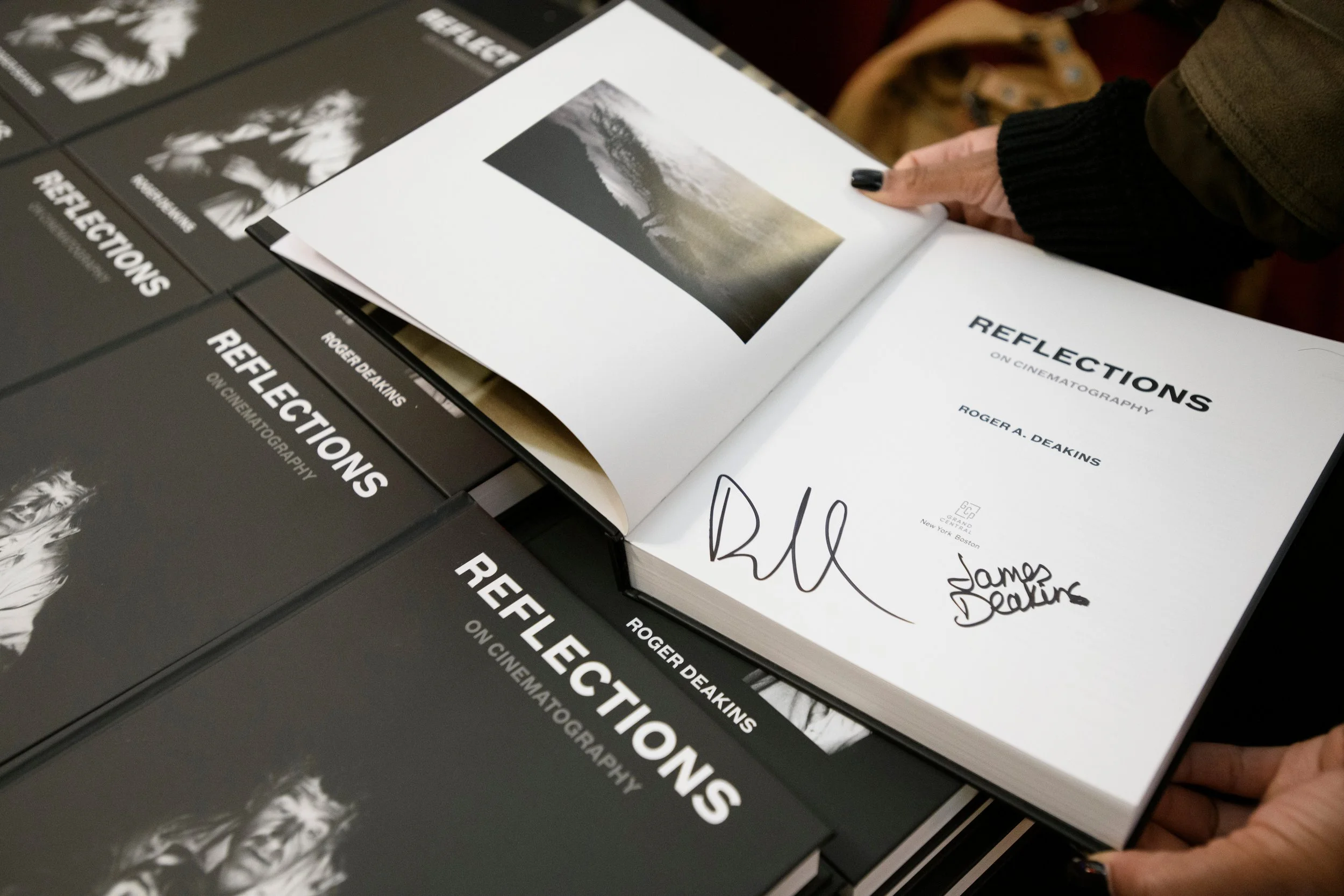 Close-up of a book titled "Reflections on Cinematography" by Roger A. Deakins, with a signature and handwritten note by James Deakin on the title page, surrounded by other copies of the same book.
