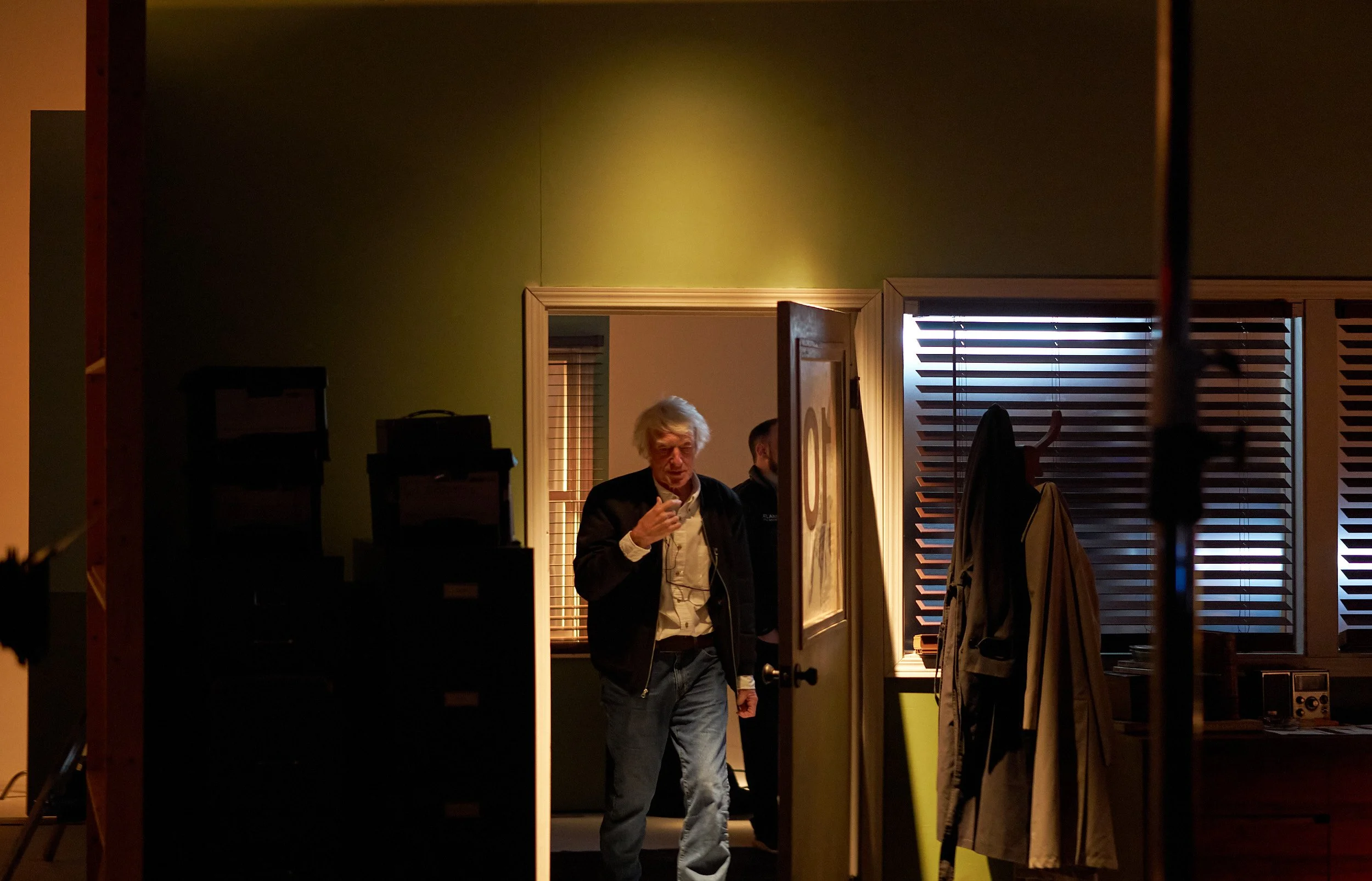 A man with white hair and a dark jacket holding eyeglasses, standing in a dimly lit room near a doorway with wood shutters on the window, and some clothing hanging on a coat rack.