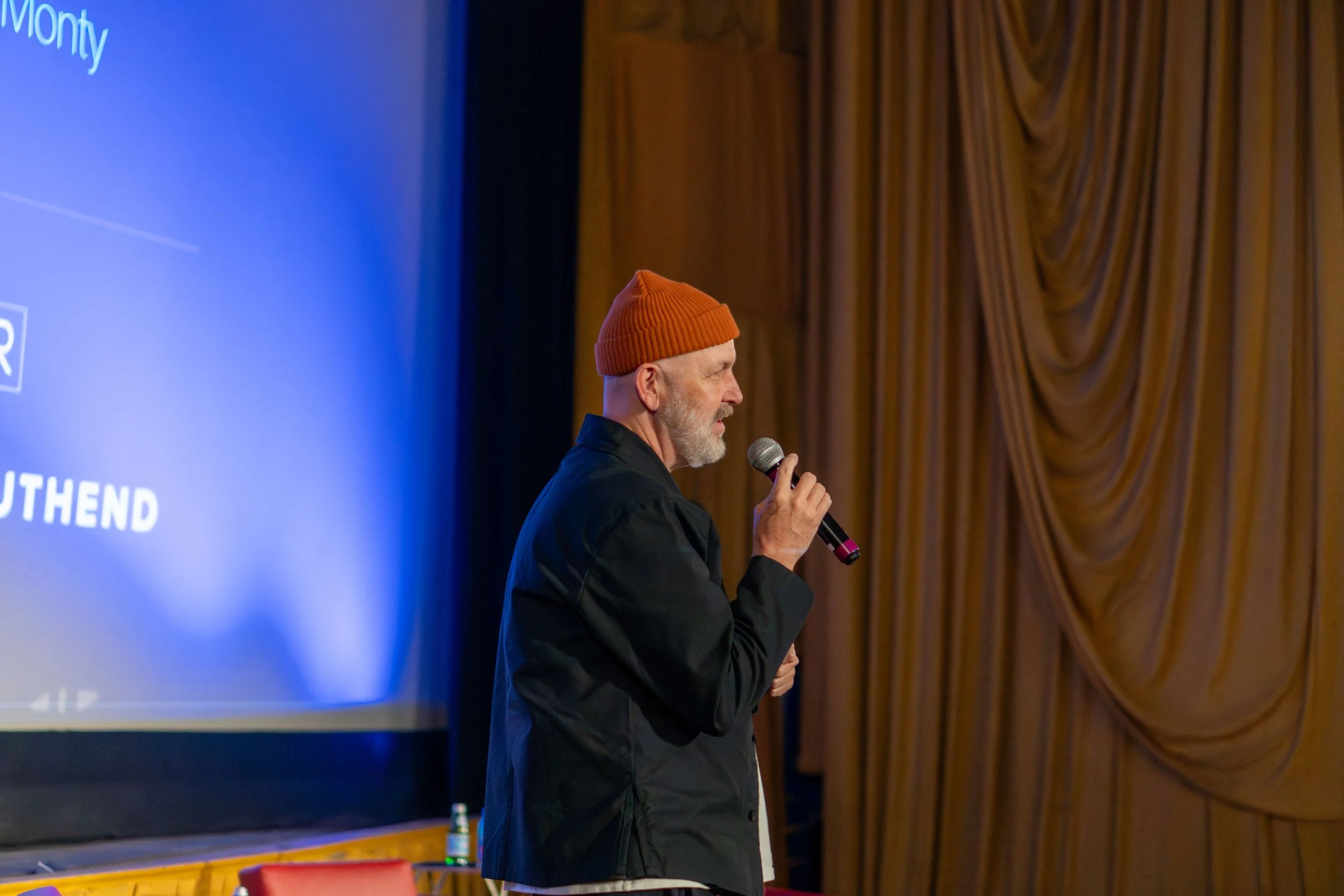 A man with a gray beard wearing an orange knit cap and black coat speaking into a microphone on stage. A large blue screen with white text is visible in the background, and gold curtains hang behind him.