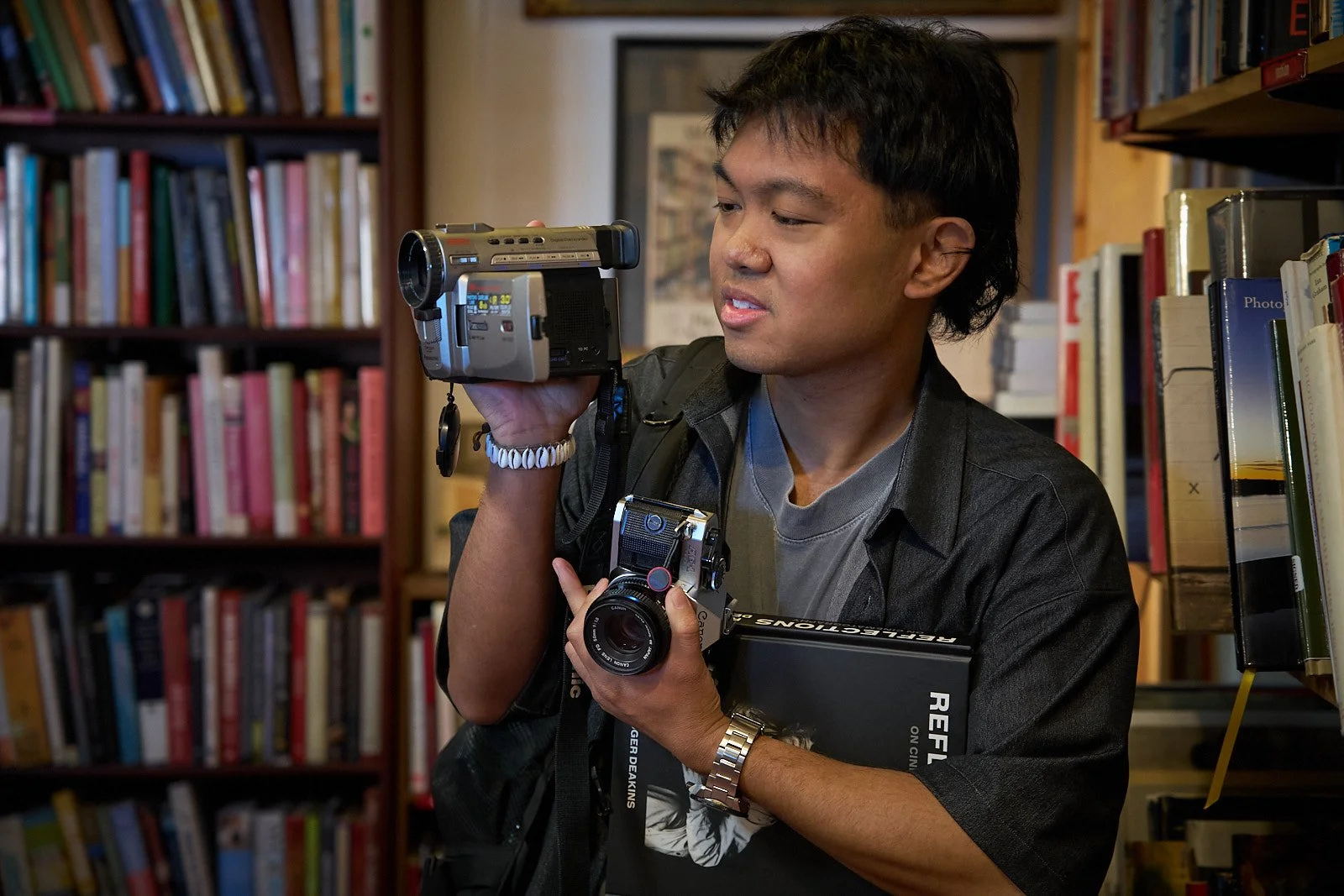 A young man in a gray shirt and black jacket in a bookstore, holding a vintage camera and a large book titled 'Reflections' while examining a video camera.