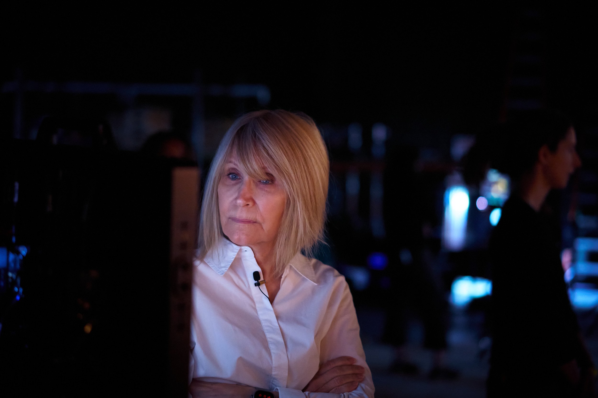 A woman with blonde hair in a white shirt standing in a dimly lit room, possibly a bar or nightclub, with blurred figures and colorful lights in the background.