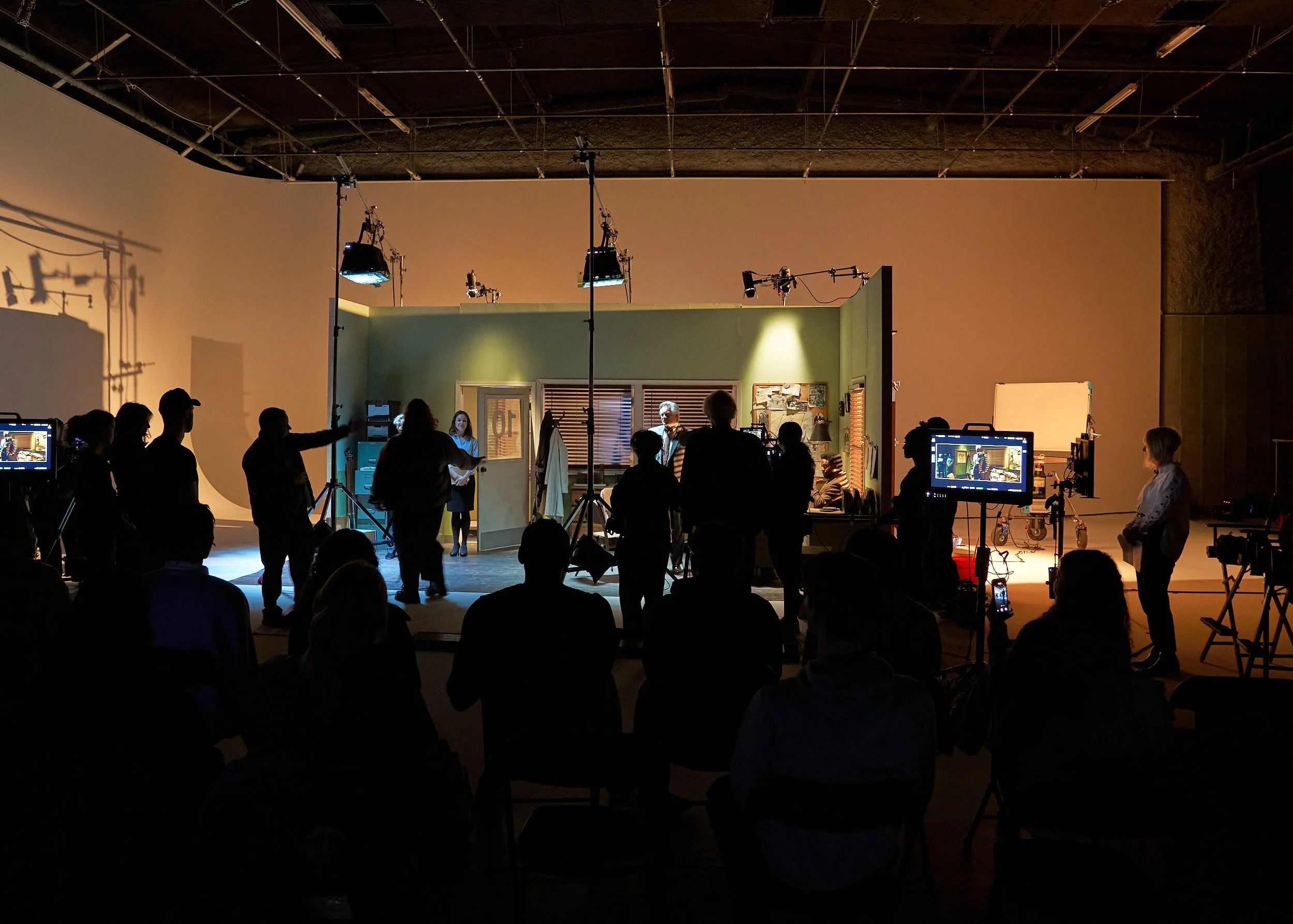 Behind the scenes of a film or TV production, with crew members working on a set with lighting, cameras, and monitors, in an indoor studio with a green backdrop and crew members discussing and preparing for a shoot.