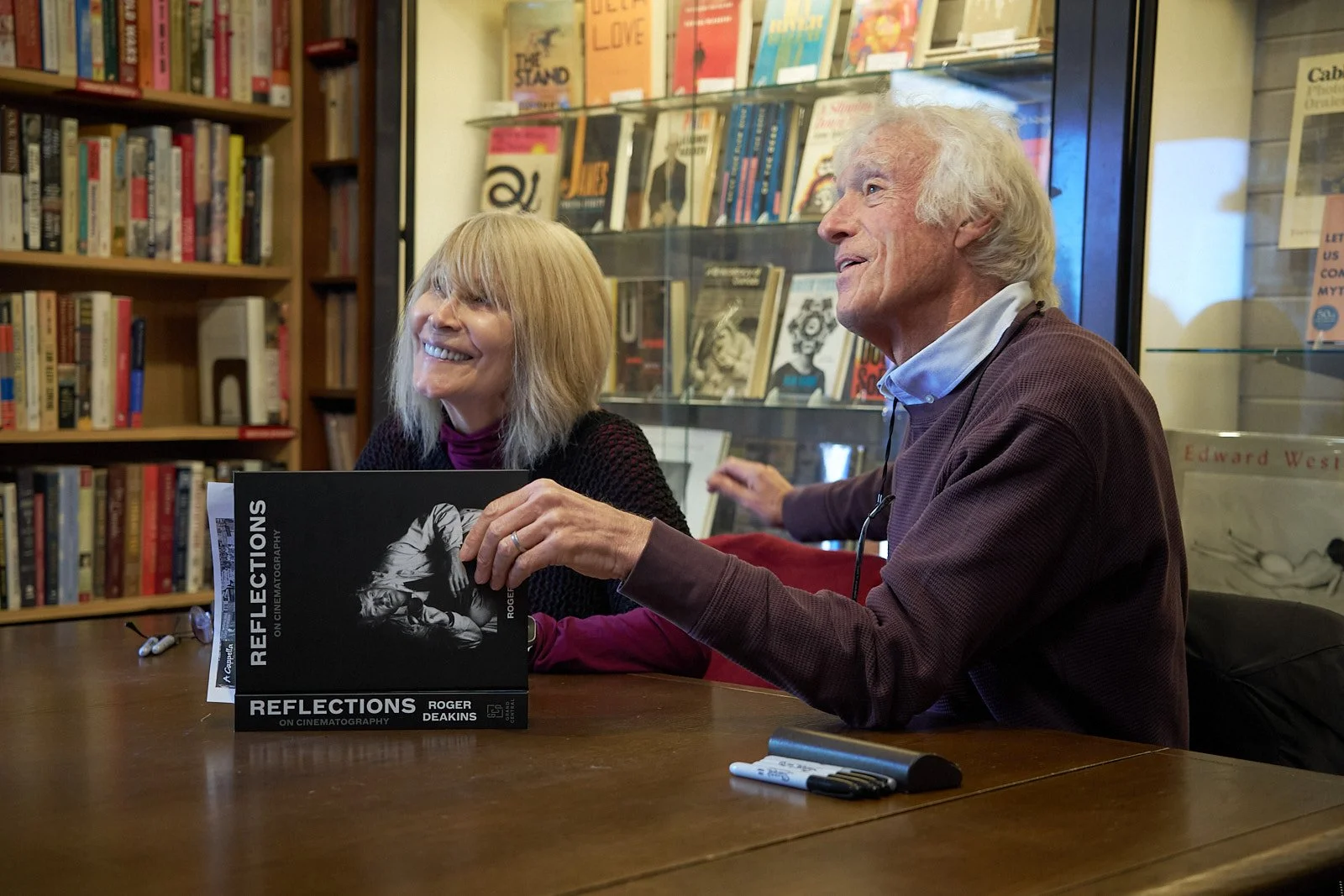 Two people, a woman and a man, sitting at a table in a bookstore or library, smiling and engaged in conversation. The woman is holding a book titled 'Reflections' by Roger Deakins, with bookshelves filled with books behind them.