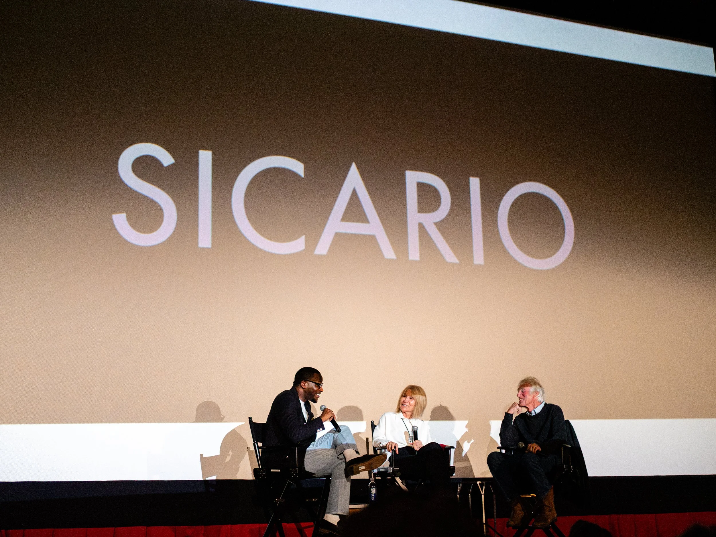 Three people sitting on stage engaged in a panel discussion. A man is speaking into a microphone, two women are listening and smiling. Behind them, a large screen displays the words 'SICARIO'.