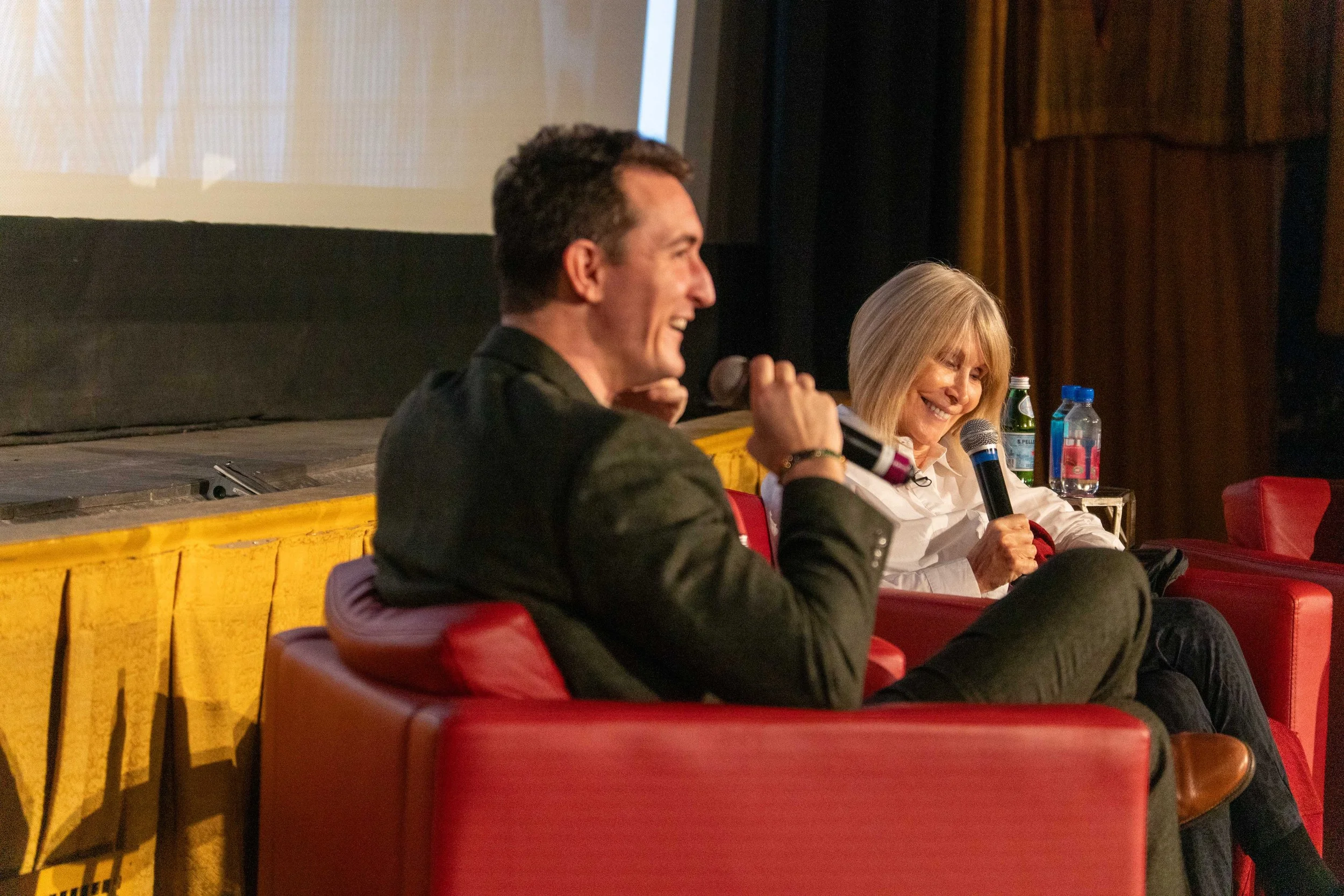 Two people seated on a stage, one man with dark hair holding a microphone, smiling, and a woman with blonde hair also smiling, holding a microphone, with water bottles on a table beside her. They are engaged in a conversation or presentation.