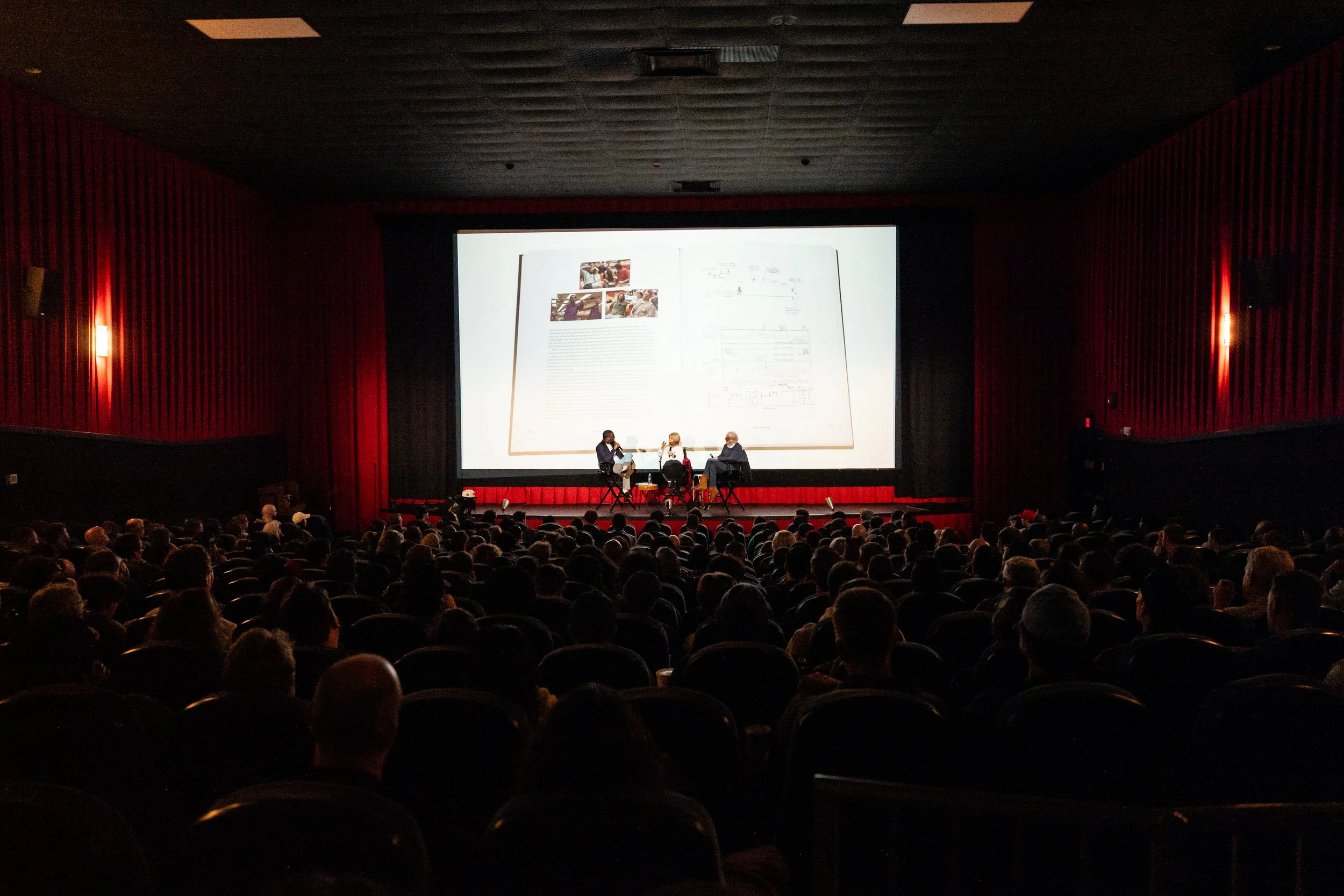 A cinema theater filled with people watching a presentation on a large screen. Three individuals are seated on stage with a large screen behind them showing a presentation slide and images. The auditorium has red walls, and the audience is seated in 