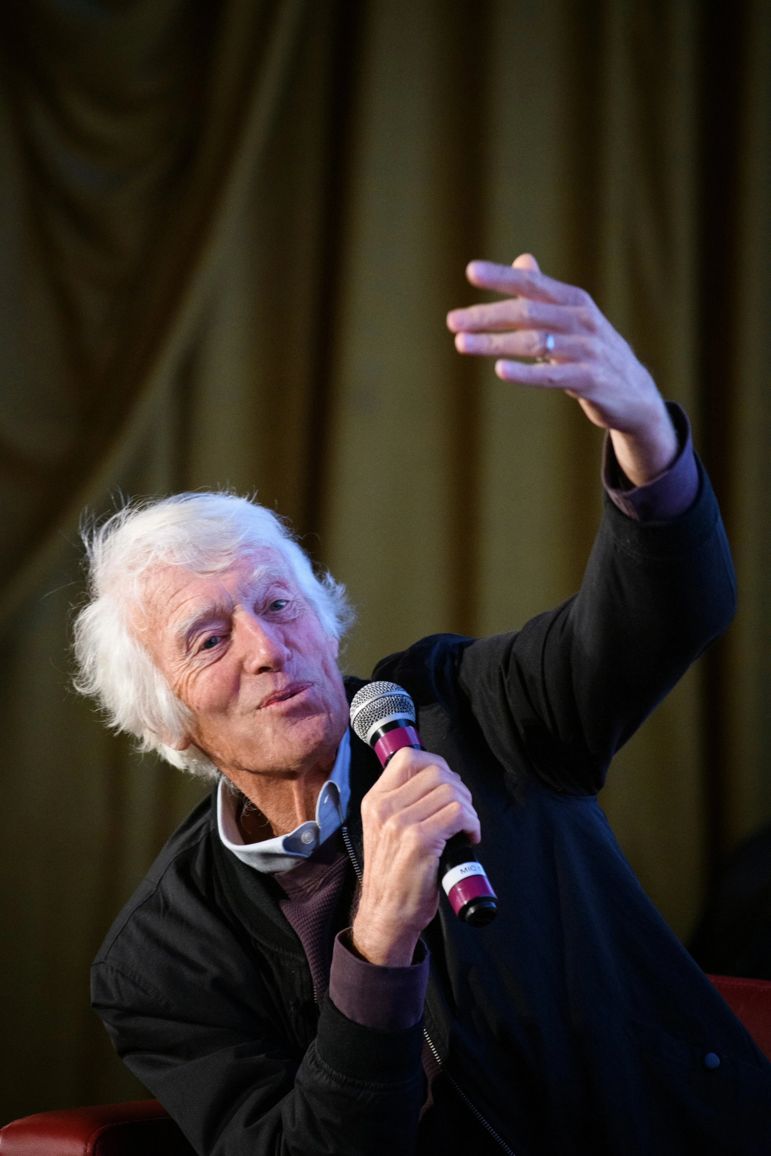 An elderly man with white hair is speaking into a microphone while gesturing with his hand. He is wearing a dark jacket and a collared shirt. The background appears to be a room with brown curtains.