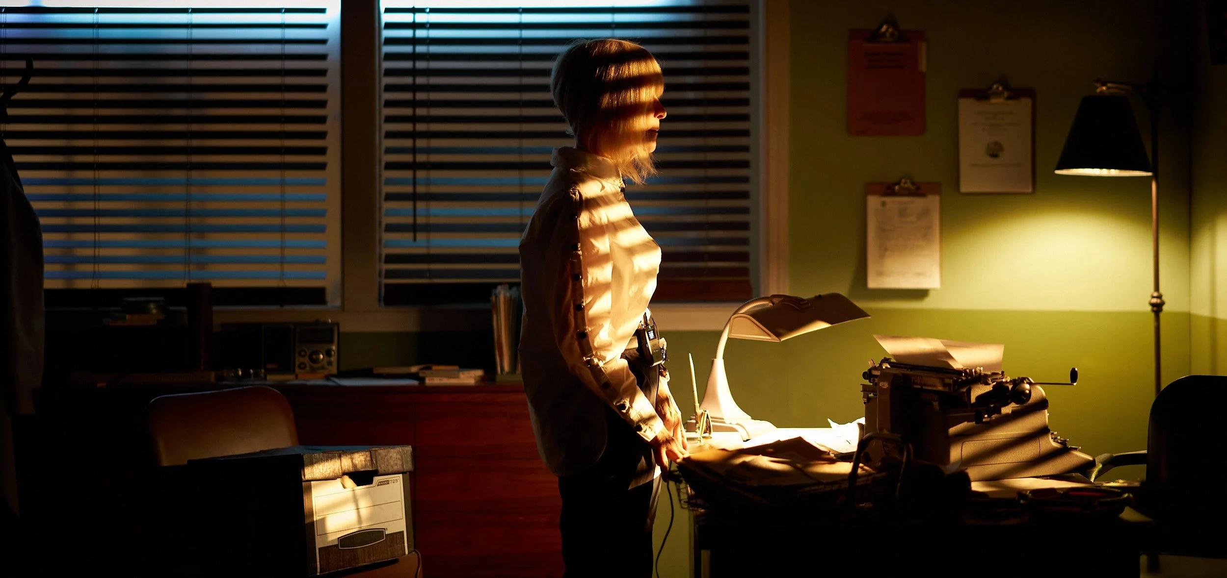 A woman standing in a dimly lit office, illuminated by the light from a desk lamp. She is looking at papers and a typewriter on the cluttered desk. The background shows window blinds and framed certificates on the wall.