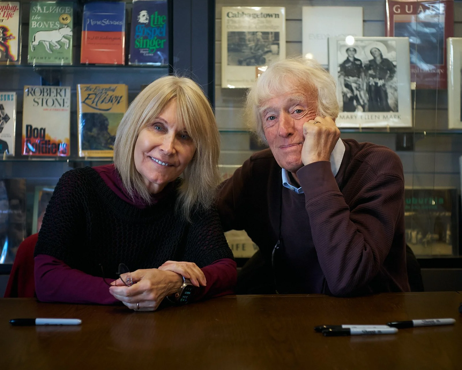 A woman and an elderly man sitting at a table in a bookstore or library, smiling at the camera. The background has bookshelves filled with books and photos.