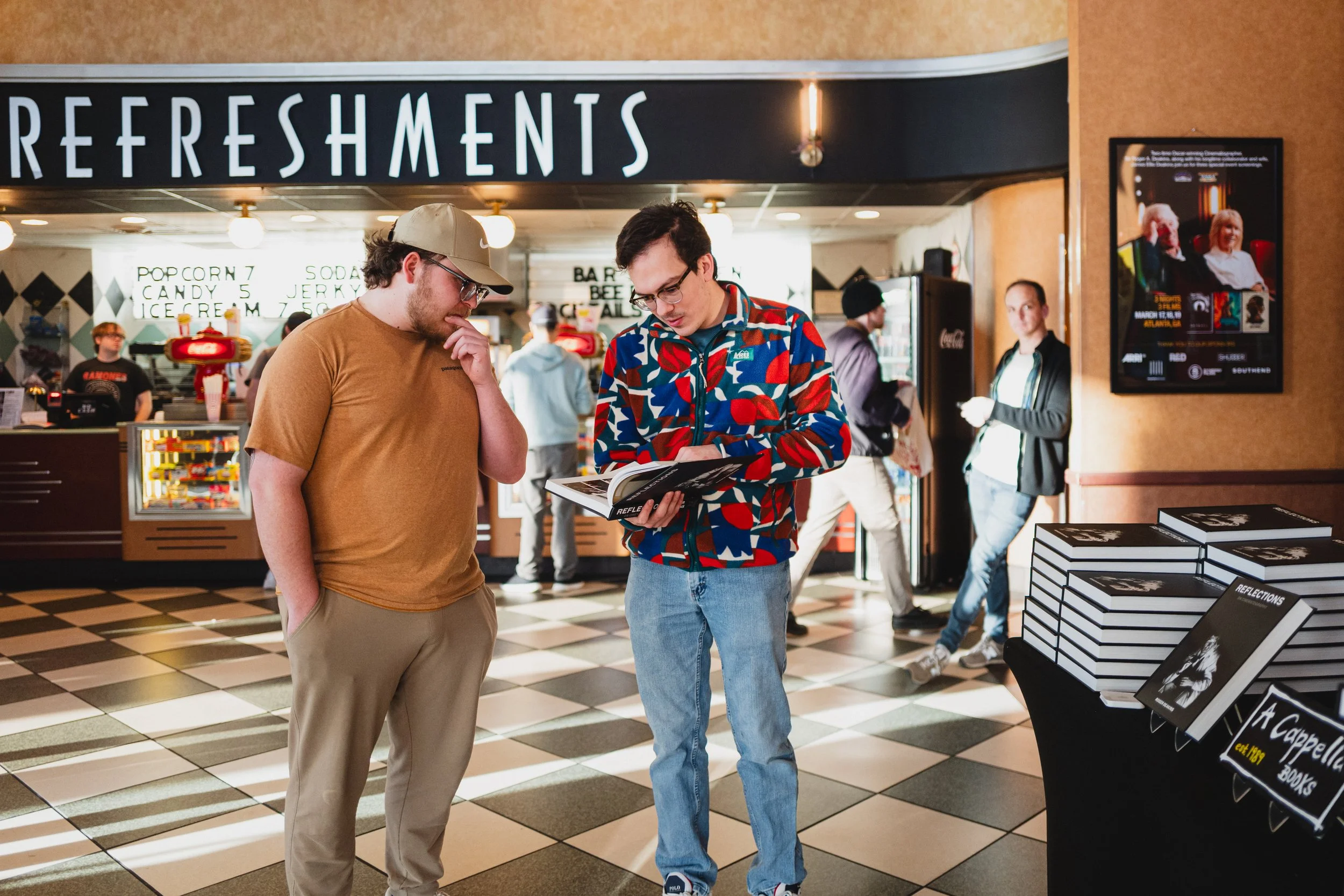 Two young men standing in a shopping mall near a souvenir book display, looking at a magazine. One is wearing a tan T-shirt and a beige cap, the other is wearing a colorful patterned jacket. In the background, there are people at the refreshment stan