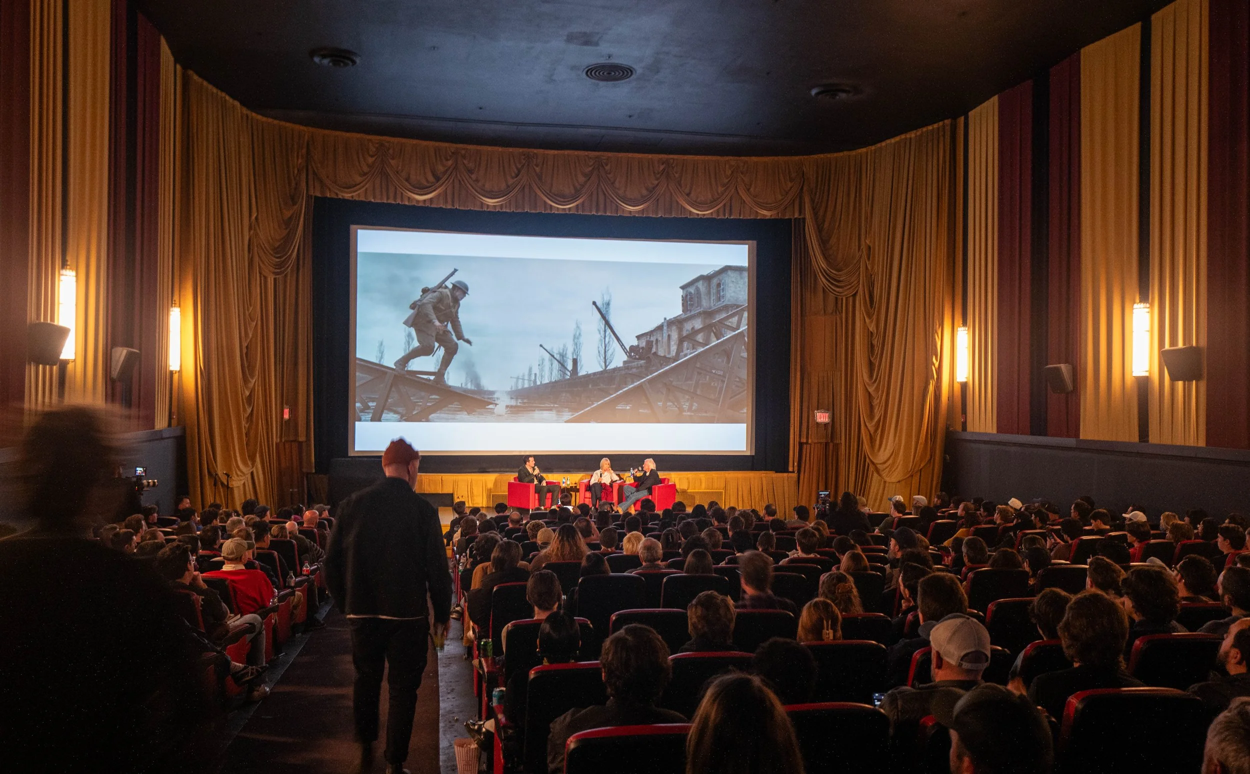 A large audience is seated in a theater watching a presentation on all three screens. The theater has gold-colored curtains and wall panels. On the main screen, a historical scene depicts soldiers wading through water and a damaged building, suggesti