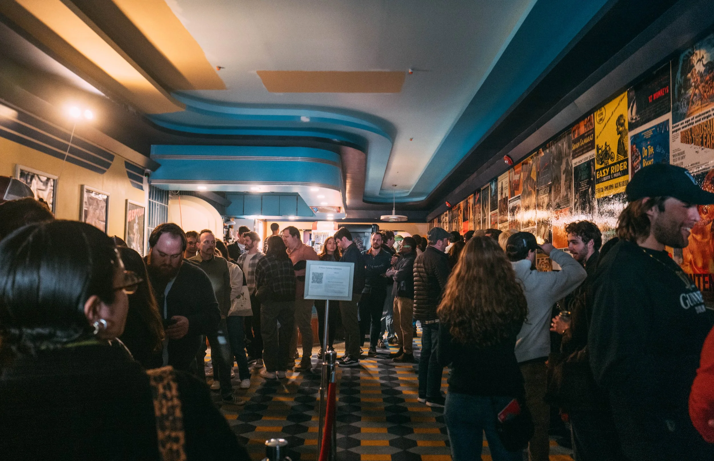 A crowded movie theater lobby with people standing in line, talking, and waiting. The ceiling has a colorful design with blue, orange, and beige shapes. The walls are decorated with posters of movies, and the floor has a geometric black, yellow, and 