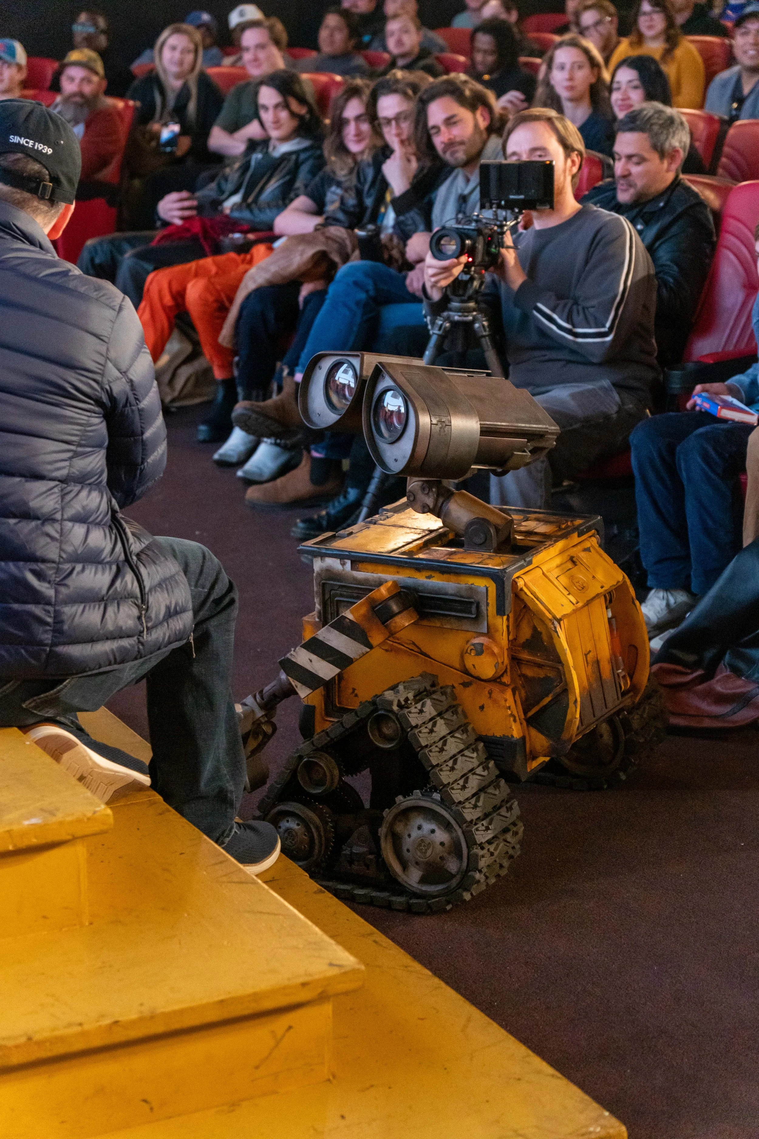 A group of people sitting in theater seats, with a person operating a camera and a toy robot resembling WALL-E in front of them. The people are smiling, and the setting appears to be a film screening or panel discussion.