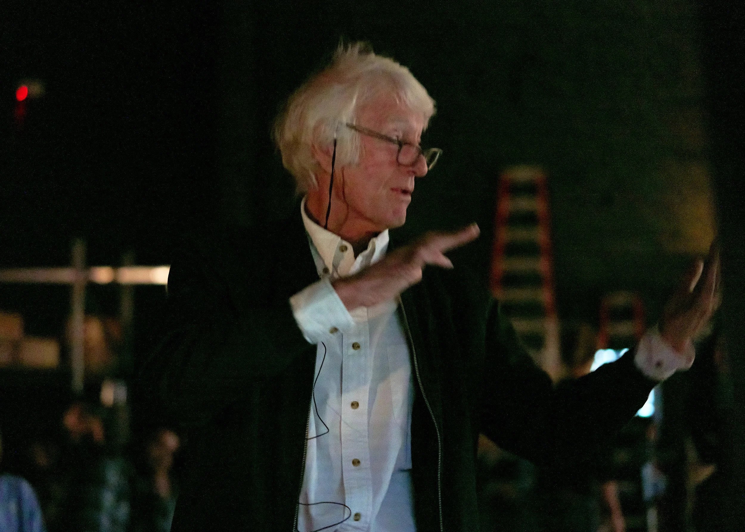 A man with white hair and glasses, wearing a white shirt and a black jacket, is speaking or making a gesture with his right hand, in a dark outdoor setting at night.