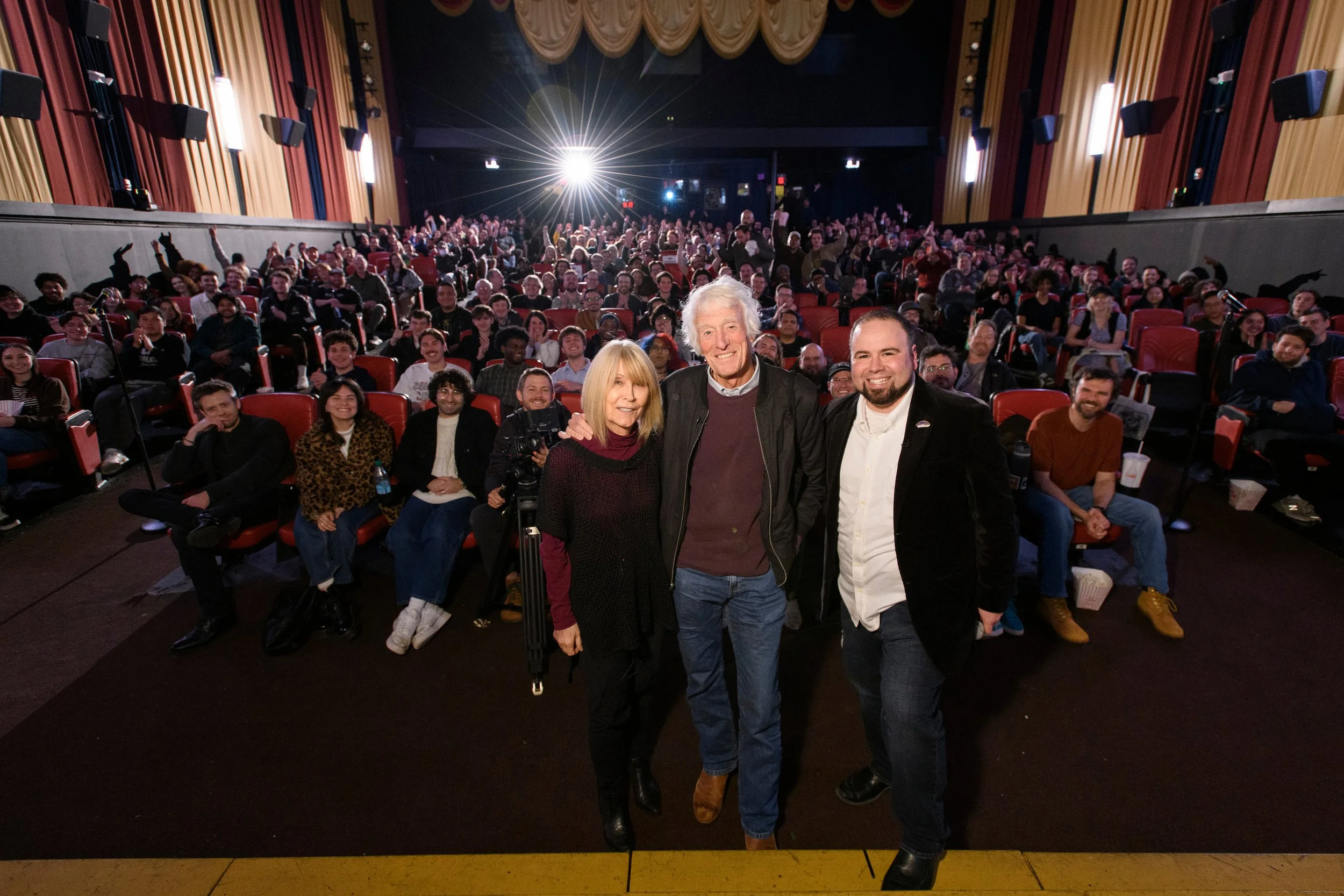 Group of people on a stage and in an auditorium, smiling for a photo, with three individuals standing at the front, two men and one woman, in an indoor theater with red and yellow curtains.