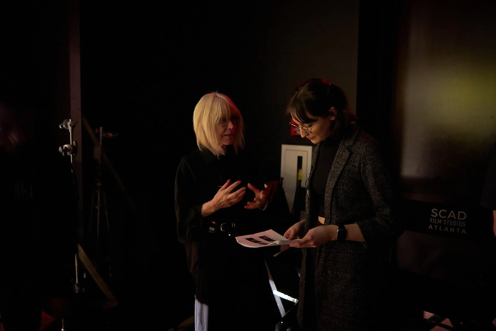 Two women standing and talking in a dimly lit room, one holding papers and showing them to the other. Background includes photography equipment and a sign that reads 'SCAD Film Studios Atlanta.'