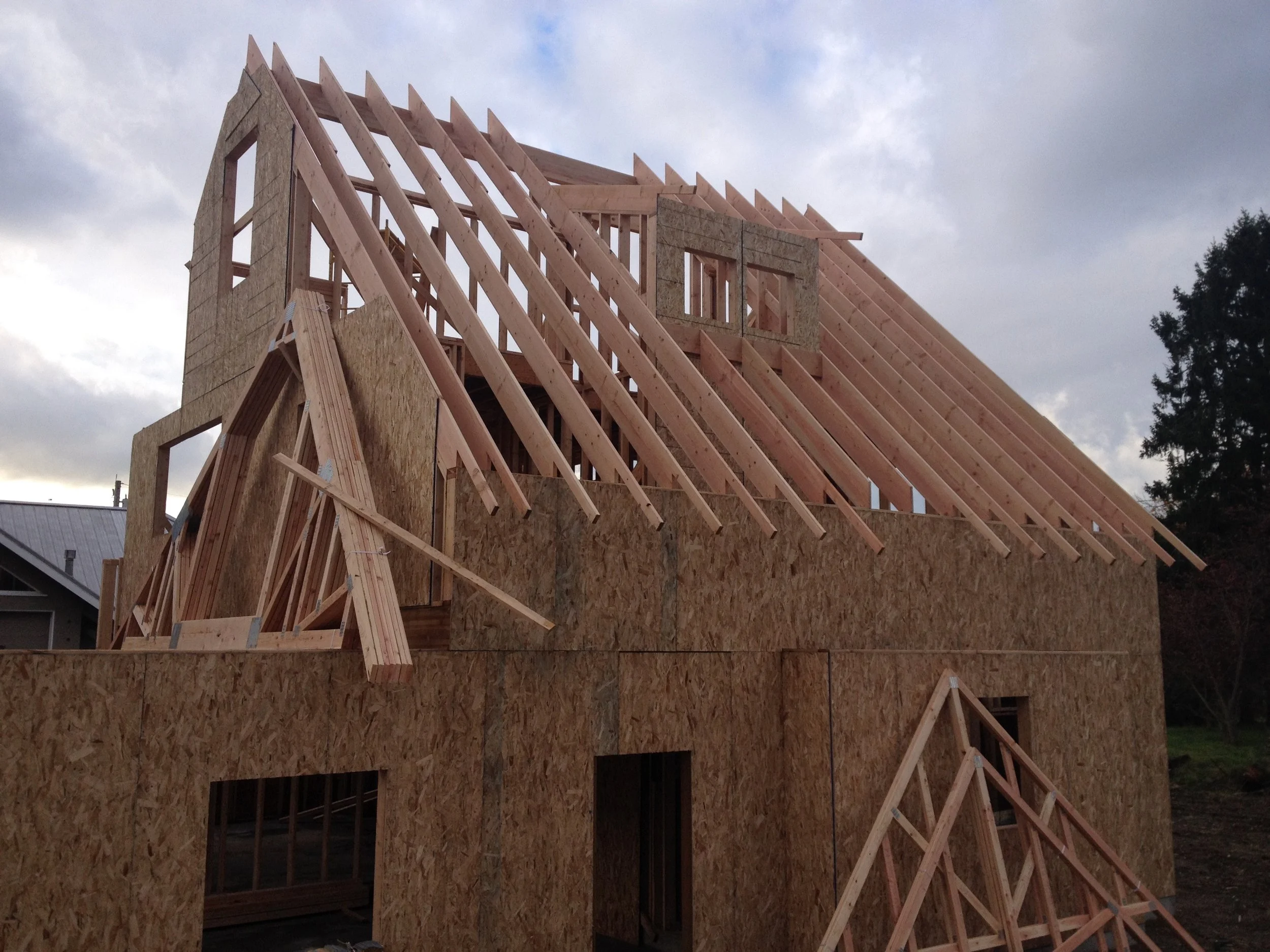 Wooden frame of a house under construction with a partly built roof and walls, with cloudy sky in the background.