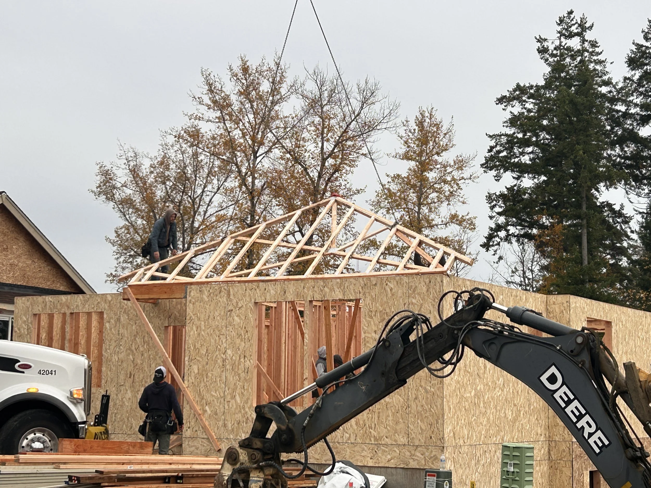 Construction workers framing the roof of a new house with wooden beams, with a large tree and cloudy sky in the background, and a construction vehicle's arm visible in the foreground.