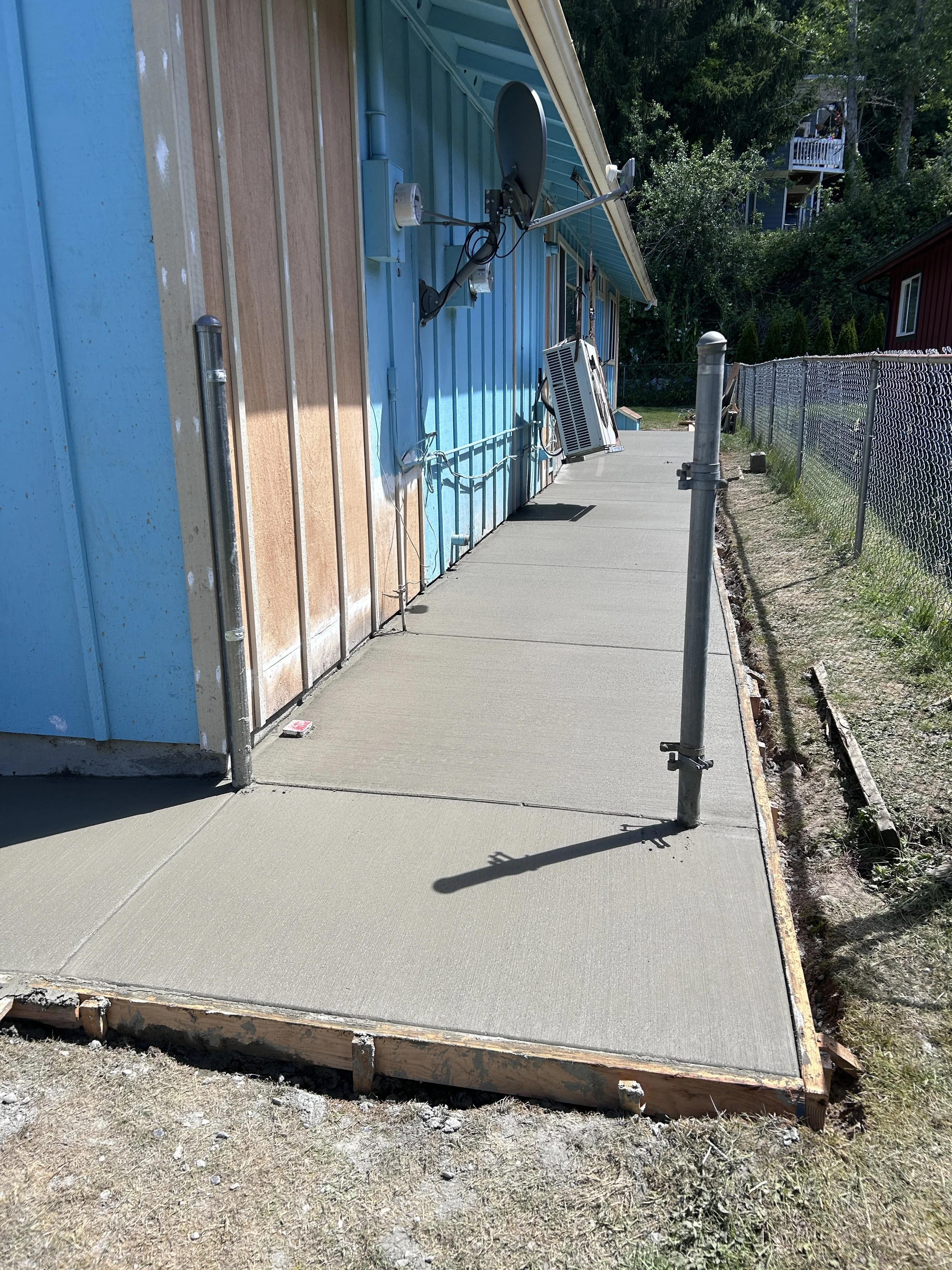 Sidewalk in front of a house with a satellite dish and air conditioning unit attached to the blue exterior wall, with a chain-link fence and trees in the background.