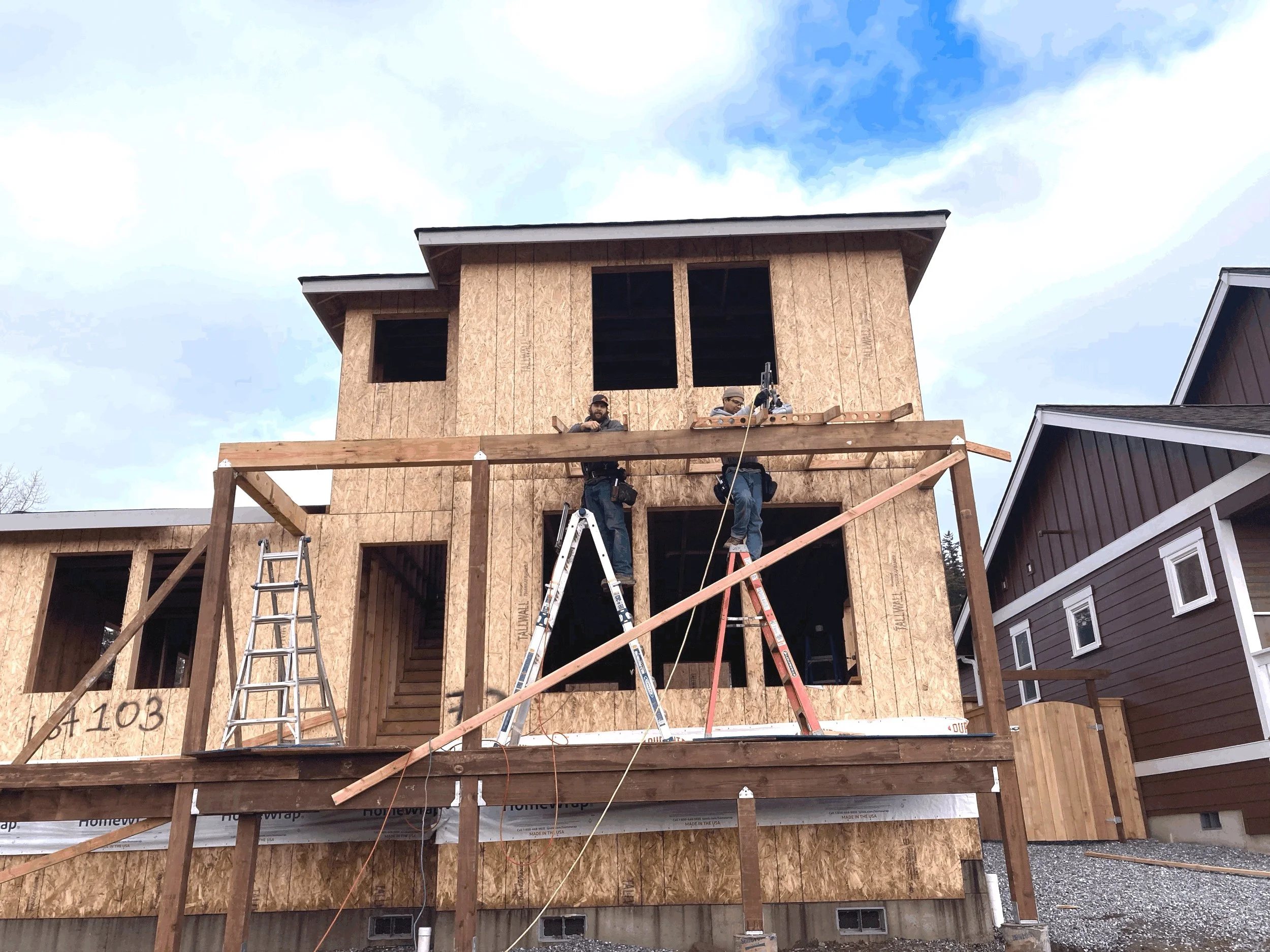 Two construction workers working on the upper floor of a house under construction, with scaffolding and ladders around them. The house has wooden framing and no windows installed yet.