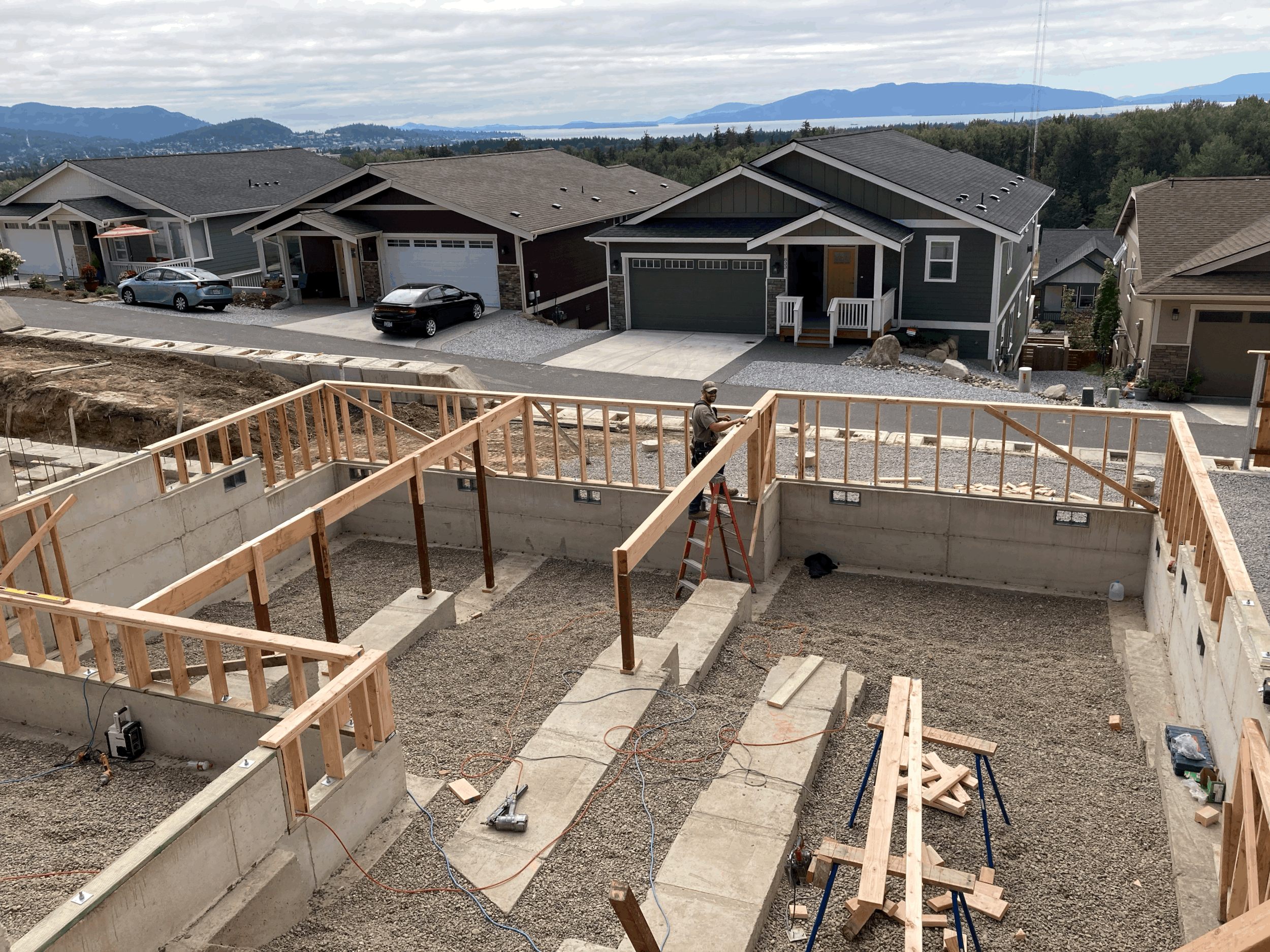 A view of a hillside from a construction site showing family houses with mountains in the background. The foreground features the foundation and framing of a new building under construction.