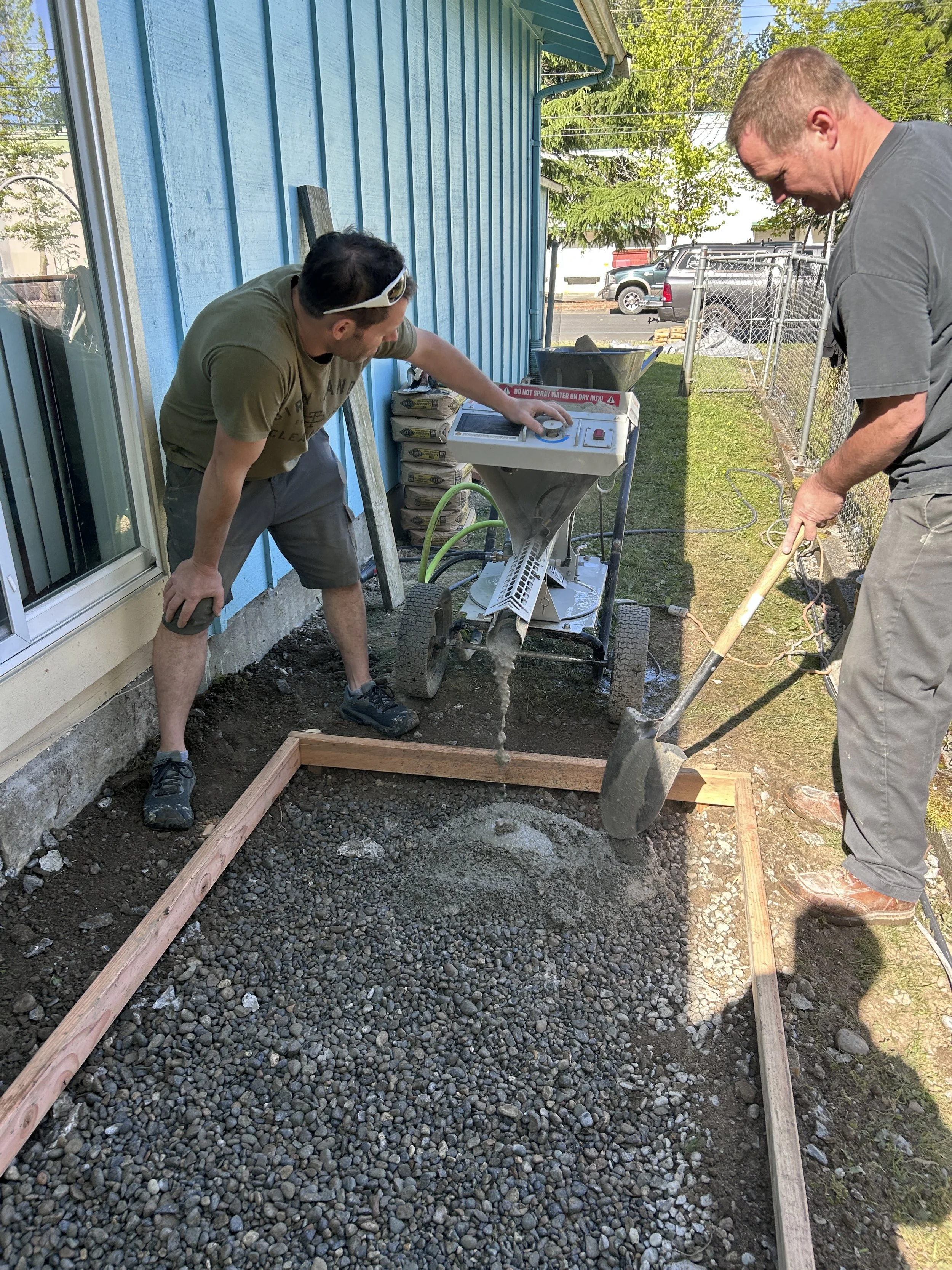 Two men working on a backyard landscaping project, spreading gravel inside a wooden frame, with tools and a concrete mixer nearby.