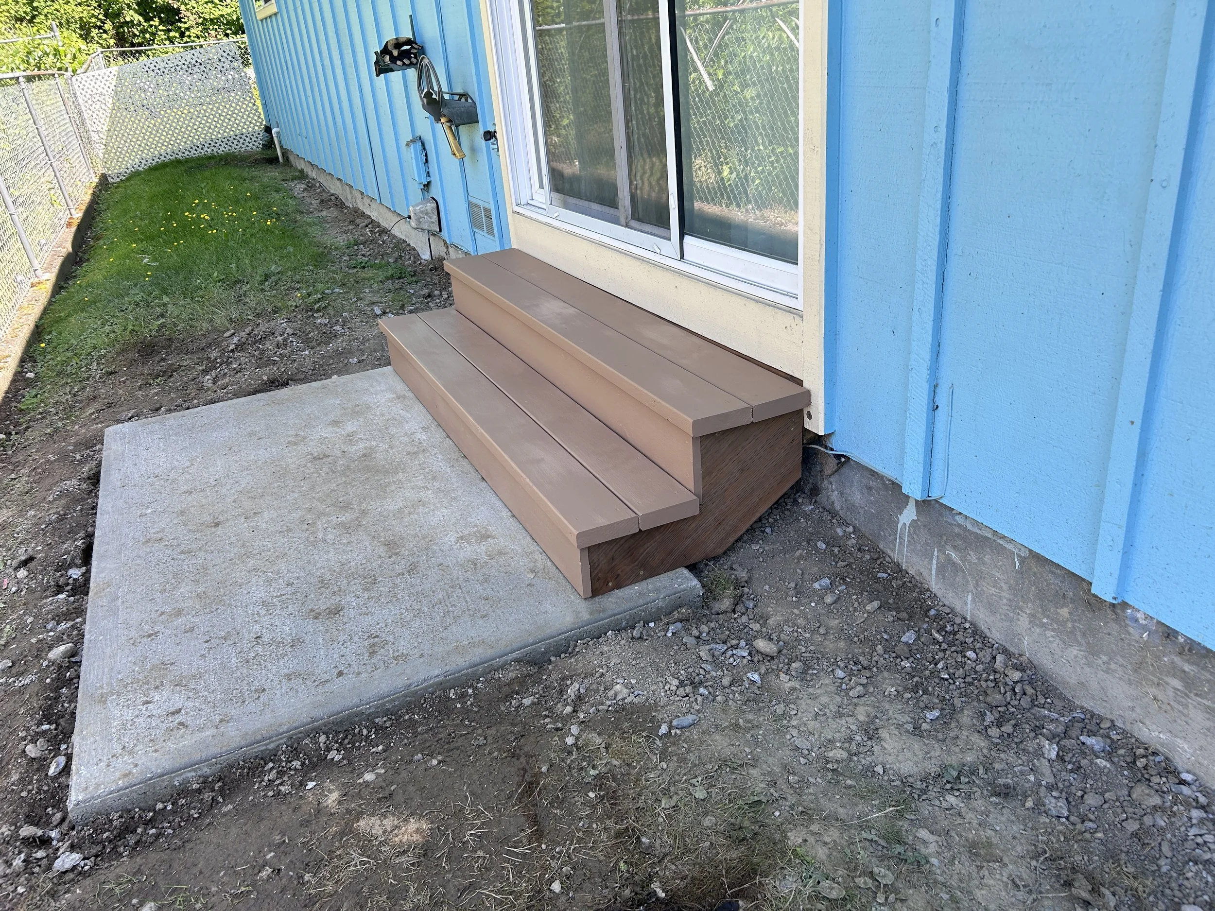 A small wooden step in front of a blue house sliding glass door with a metal lock. The area outside has concrete, dirt, grass, and a chain-link fence with white lattice privacy panels.